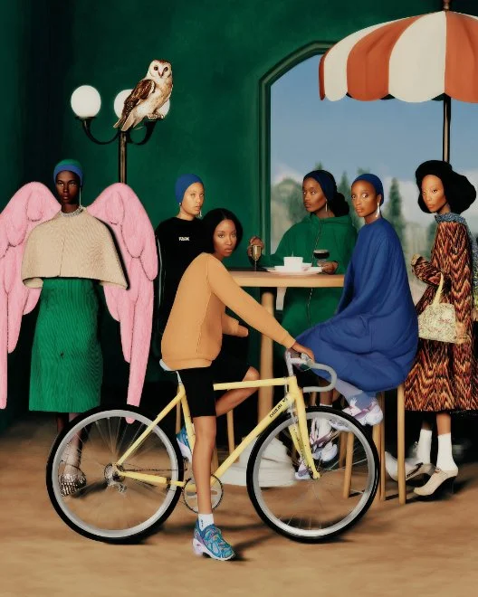 A group of six mannequins dressed in colorful clothing inside a room with a green wall, a large window, and a striped umbrella. One mannequin has pink angel wings and an owl lamp on its shoulder. A woman in a peach sweater and black shorts stands next to a yellow bicycle, and a woman in a blue dress sits at a table. Another woman in a dark green dress and a woman in a patterned dress stand nearby.