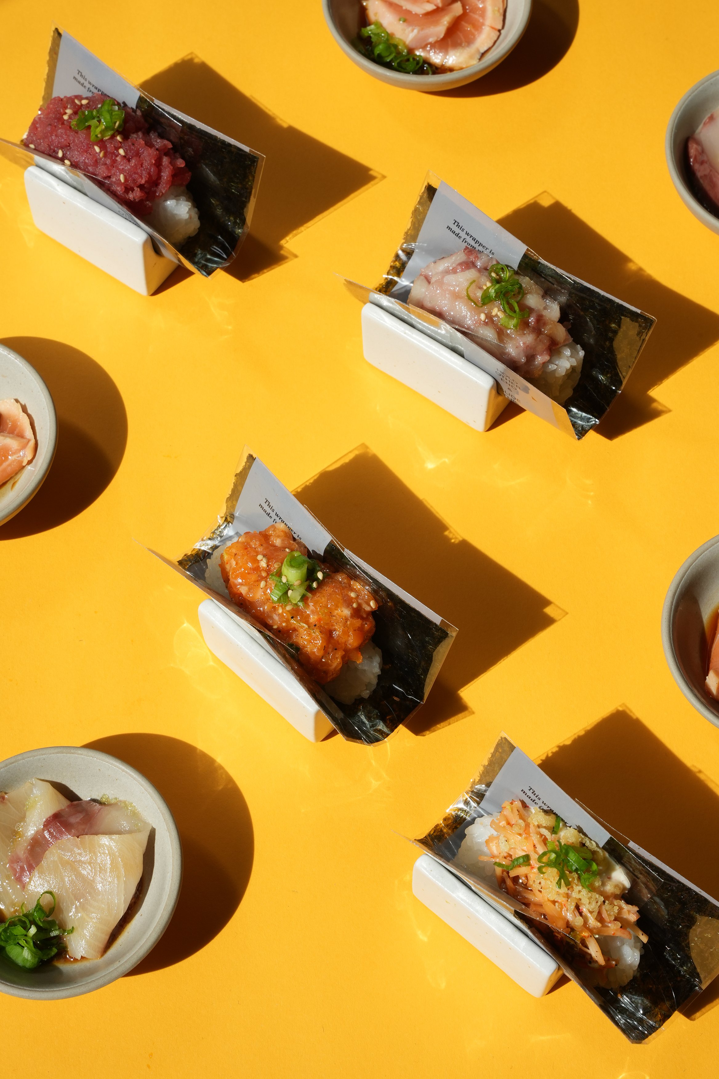Various pieces of sushi and sashimi on yellow surface, some in plastic trays and some in small bowls, with shadows cast across the surface.
