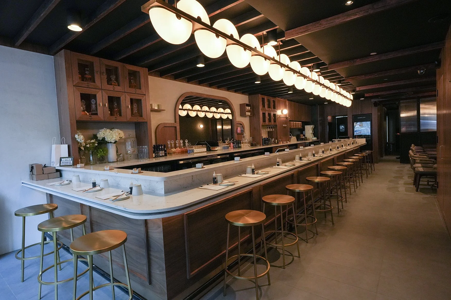 Interior of a modern bar with a long marble counter, wooden barstools, and warm lighting. The ceiling has dark beams and large round pendant lights. There are cabinets with glassware and decorative items behind the bar.