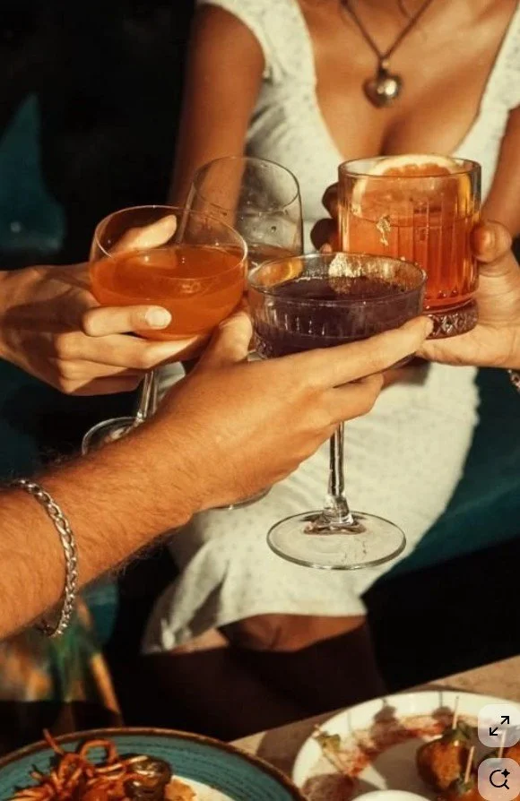 Close-up of several people clinking glasses filled with colorful cocktails at a social gathering, with a woman in a white dress visible in the background.