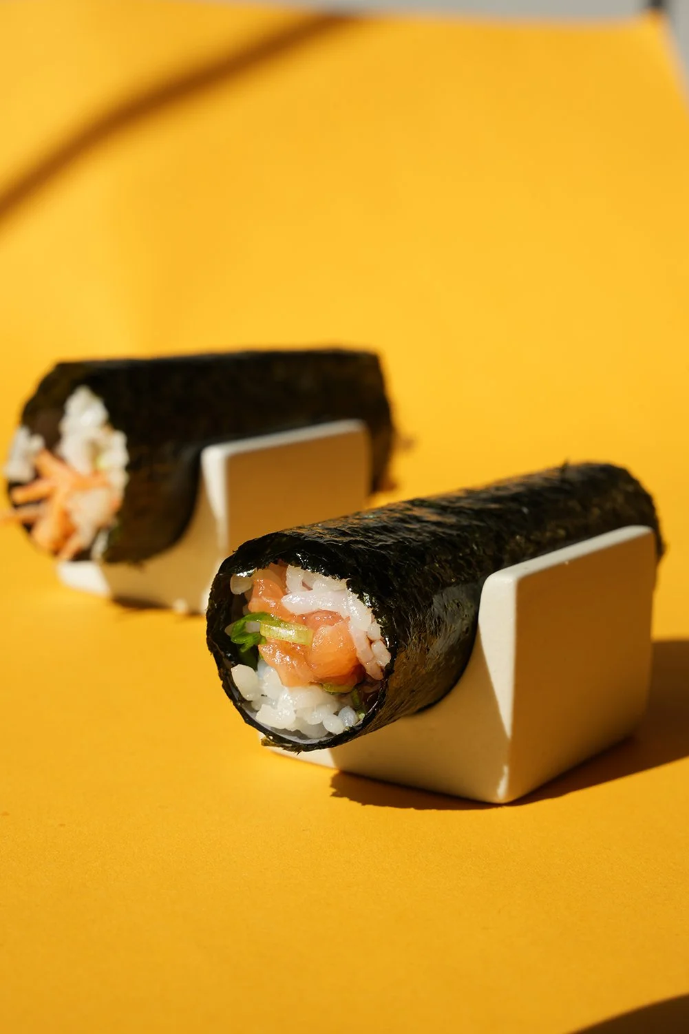 Close-up of two sushi rolls with nori seaweed wrapping, displaying salmon, rice, and green onion, placed on white holders against a yellow background.