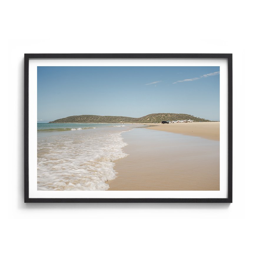 Framed coastal photograph with soft sand, gentle waves and distant headland framed in black