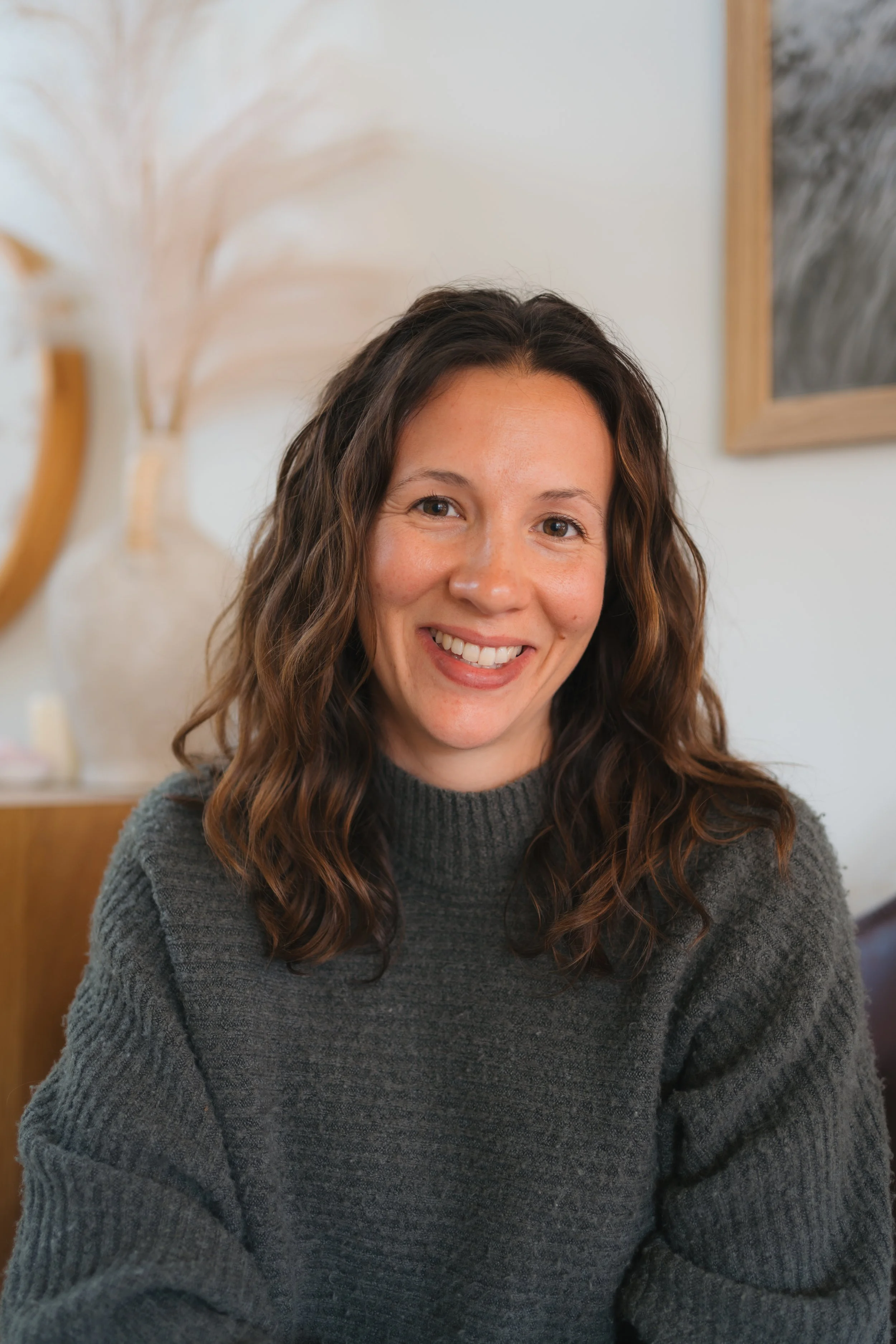 A woman with shoulder-length wavy brown hair and a warm smile, sitting indoors wearing a dark gray knitted sweater. Behind her is a blurred background with artwork and a decorative plant.