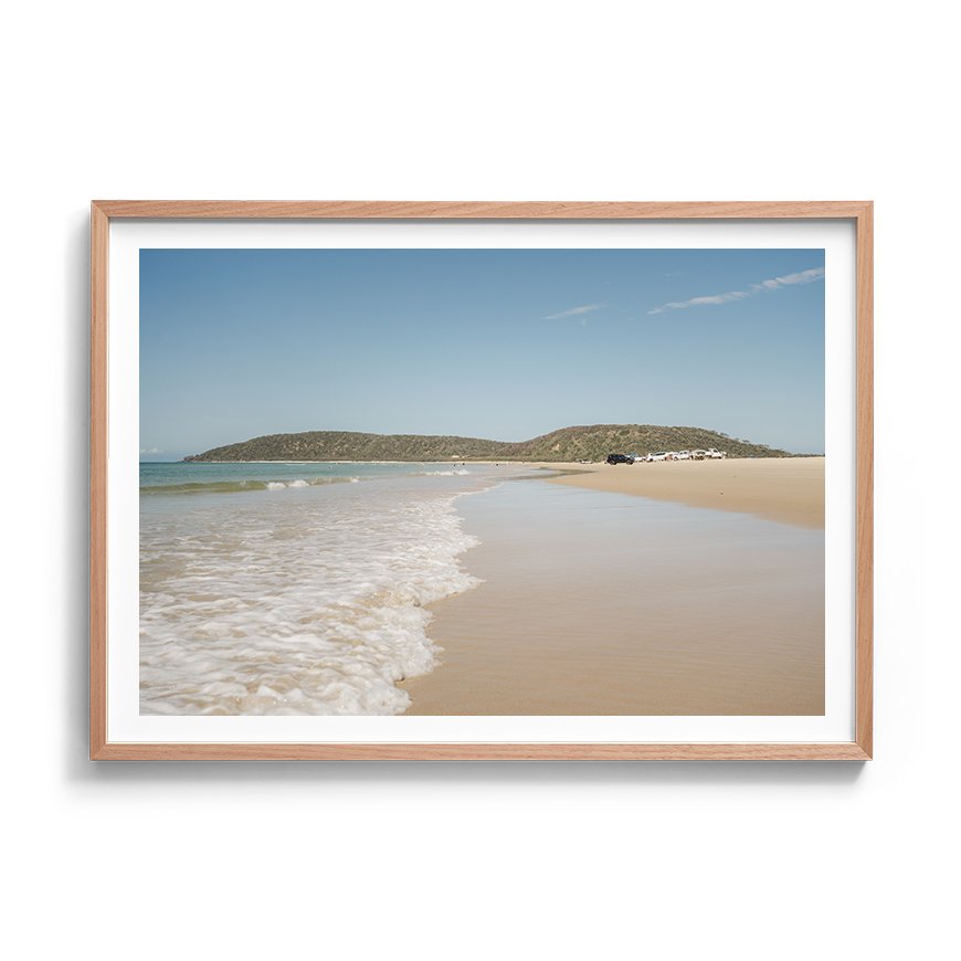 Framed coastal photograph with soft sand, gentle waves and distant headland framed in oak
