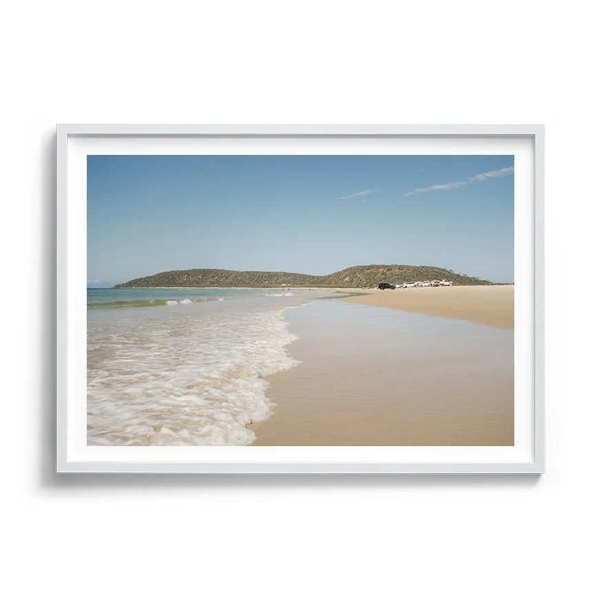 Framed coastal photograph with soft sand, gentle waves and distant headland framed in white