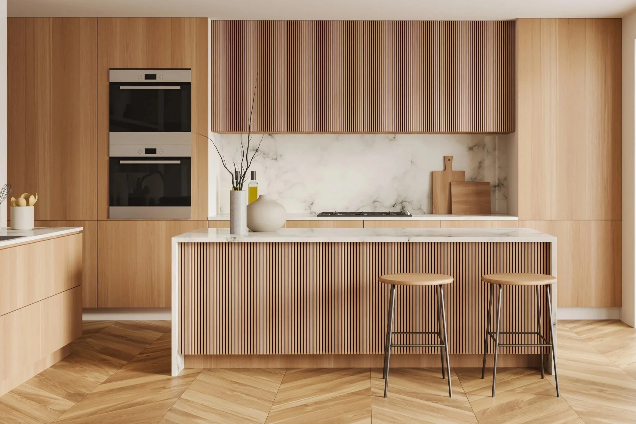 Modern kitchen with wooden cabinets, a marble countertop, and a matching marble backsplash. There are two wooden bar stools at an island, a vase with branches, a bottle of olive oil, and cutting boards on the counter. The floor has a herringbone wood pattern.