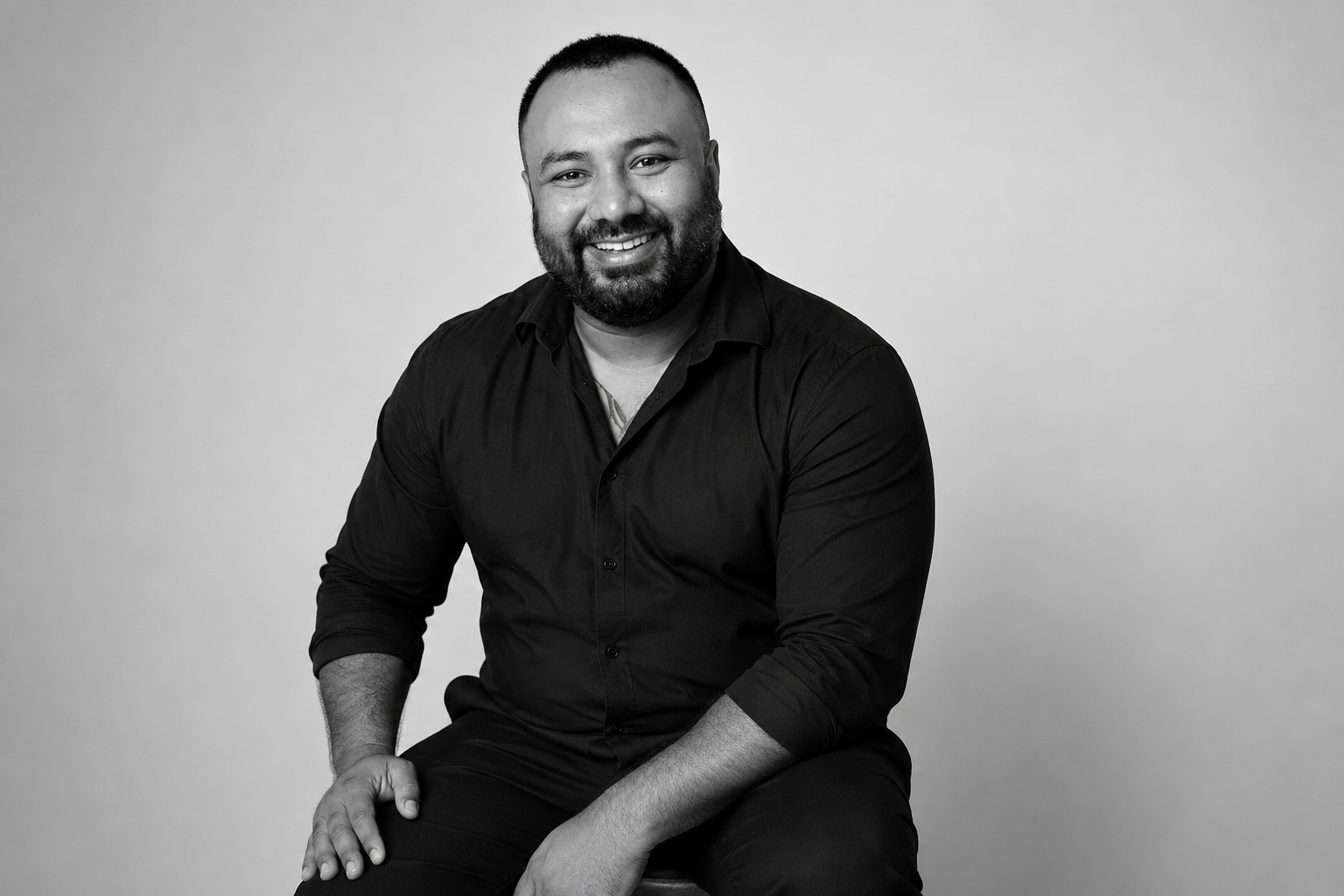 Black and white studio portrait of a smiling man with short hair and beard, wearing a black button-up shirt, sitting on a stool with one hand resting on his knee, against a plain light gray background.