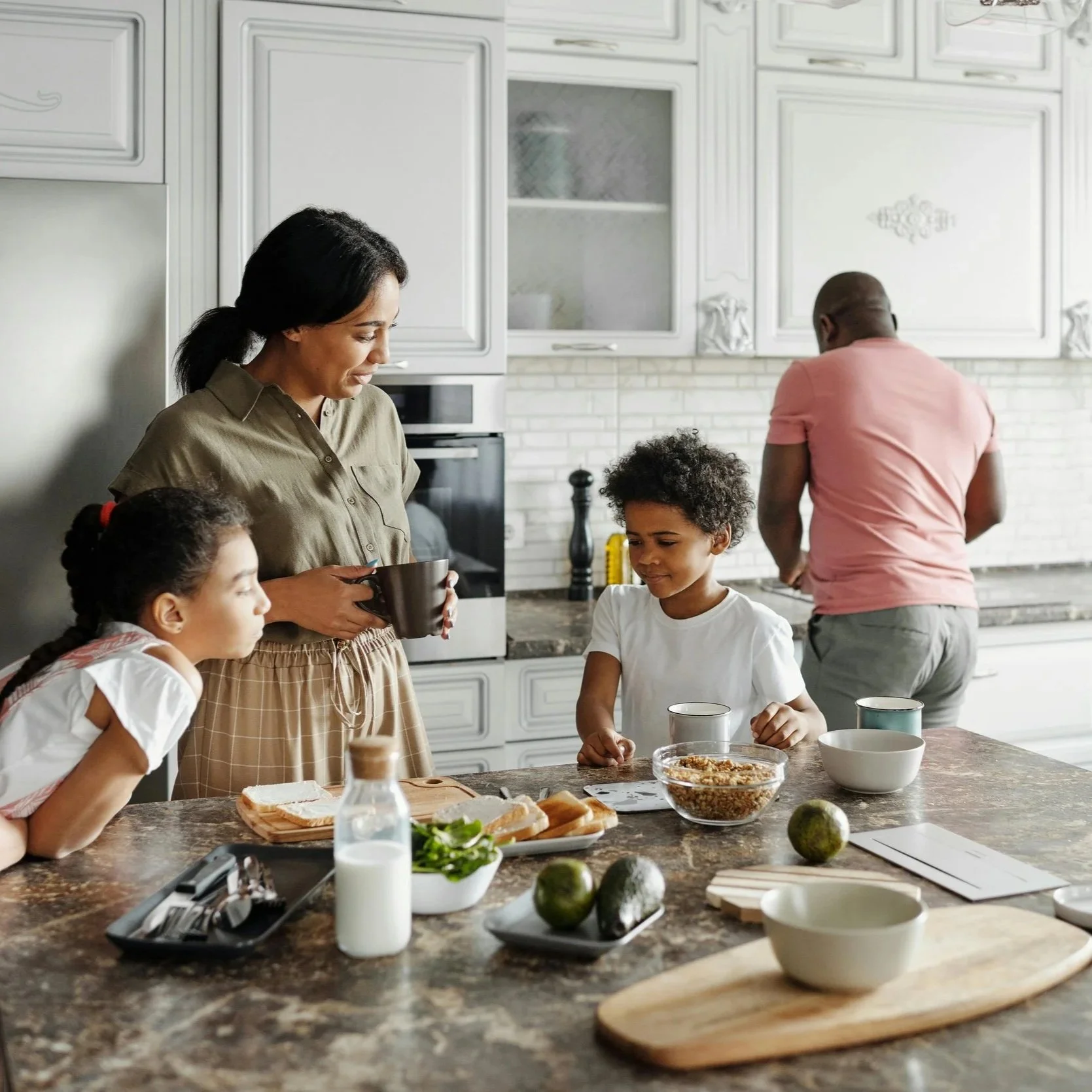 A multi-ethnic family having breakfast in a modern kitchen. Children and adults are gathered around a kitchen island with various food items and drinks, enjoying a meal together.