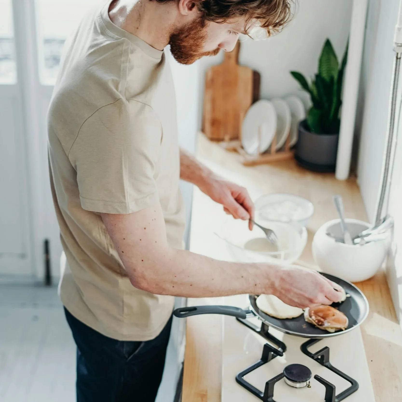 A man with a beard cooking breakfast on a stove, flipping pancakes in a frying pan in a bright kitchen.