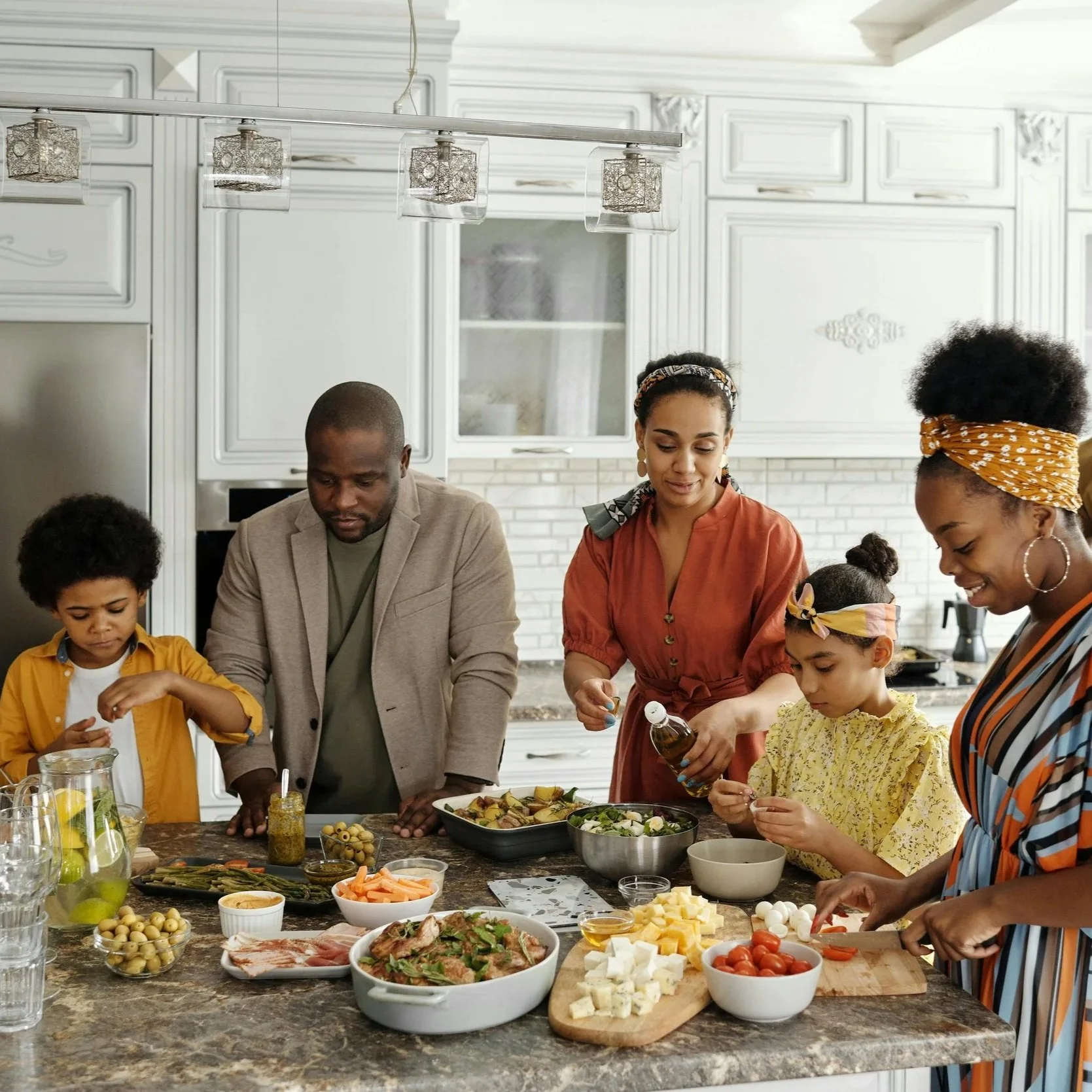 People preparing a meal together in a modern kitchen with white cabinets and a marble countertop. Various ingredients, including cheese, vegetables, and olives, are arranged on the counter.
