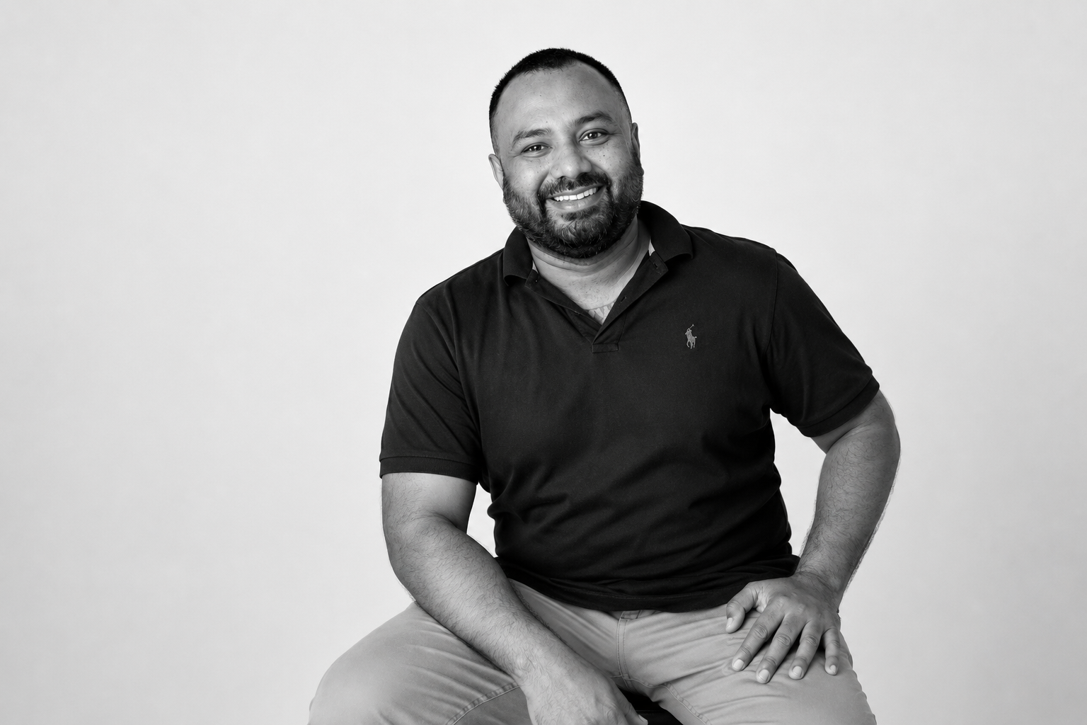 Black and white portrait of a smiling man with short hair and a beard, wearing a black polo shirt and light-colored pants, sitting against a plain background.