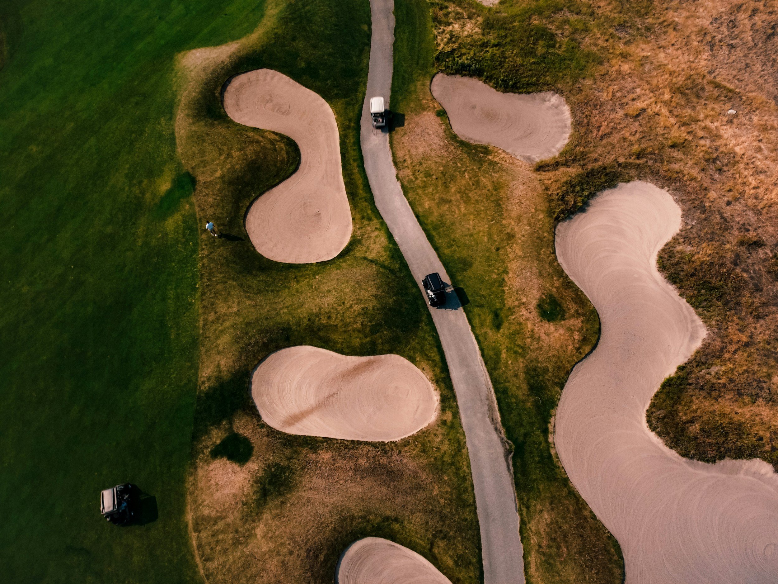 An aerial view of a golf course with multiple sand bunkers and a narrow paved pathway. A person wearing a blue shirt and white cap is walking near a golf cart, while another golf cart is parked on the grass.