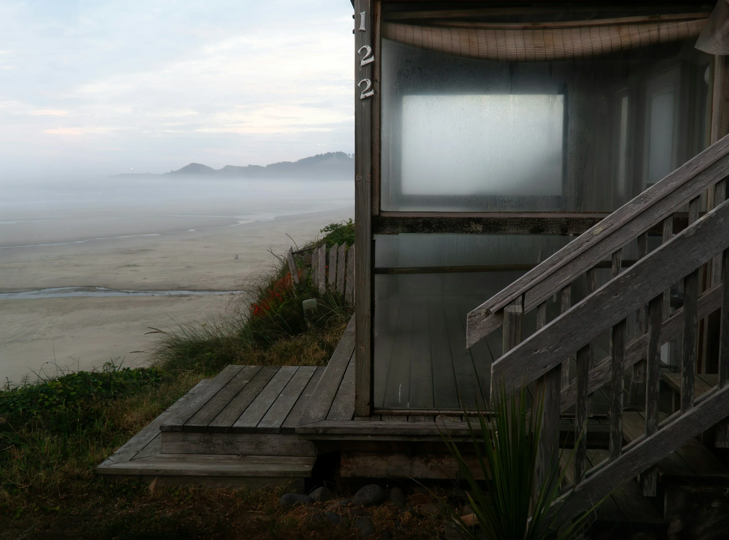 A foggy beach view with calm ocean waves, distant hills, and sandy shoreline, viewed from a wooden staircase outside a small structure with a glass window and a bamboo roof.