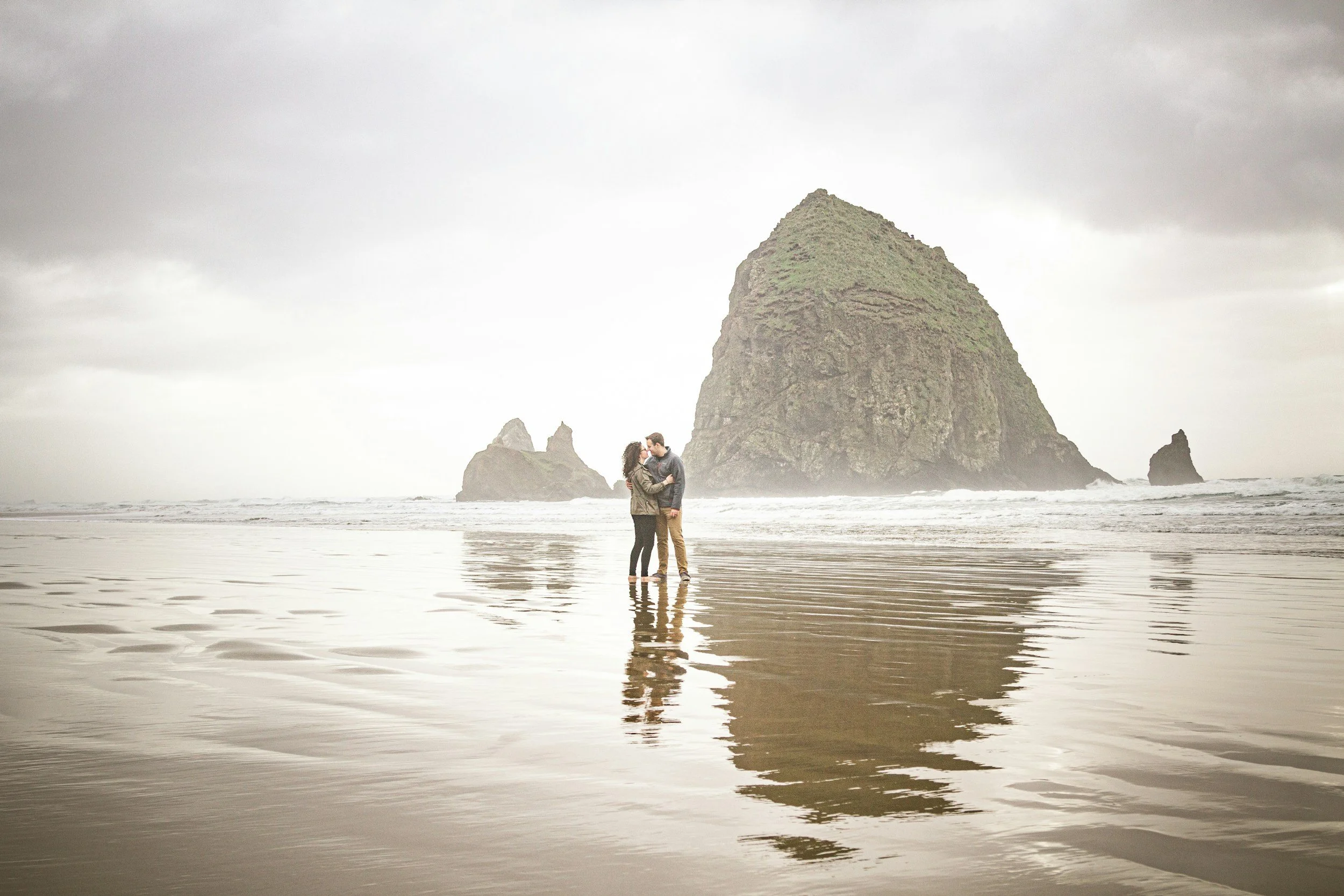 A couple stands in the shallow ocean water holding each other in front of a large rock formation on a cloudy day.