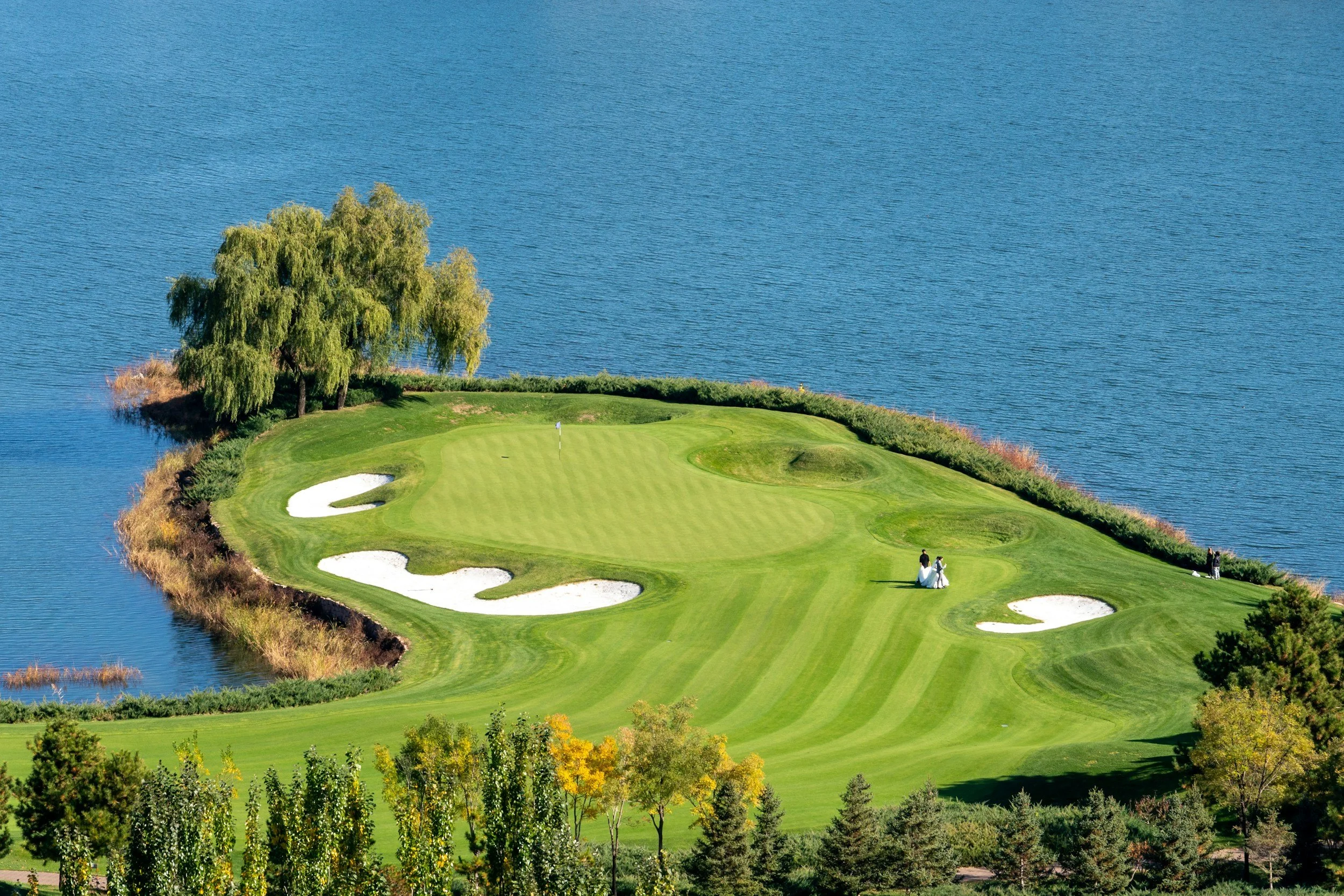 A golf course surrounded by water with a green and sand bunkers. Two people are walking near the green, and trees are visible in the background.