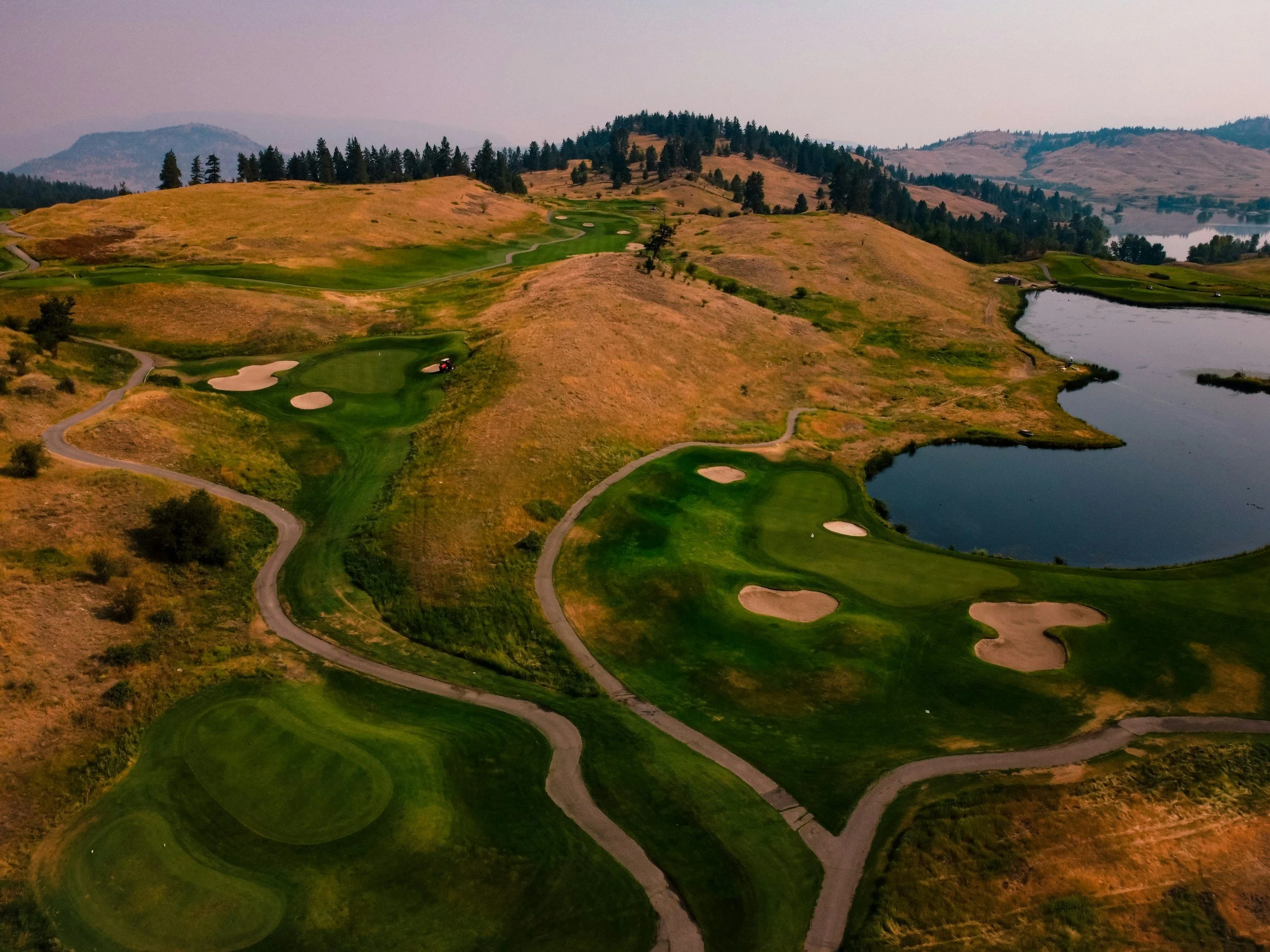 A scenic golf course with green fairways and sandy bunkers, surrounded by rolling hills and trees, with a body of water on the right side of the image.