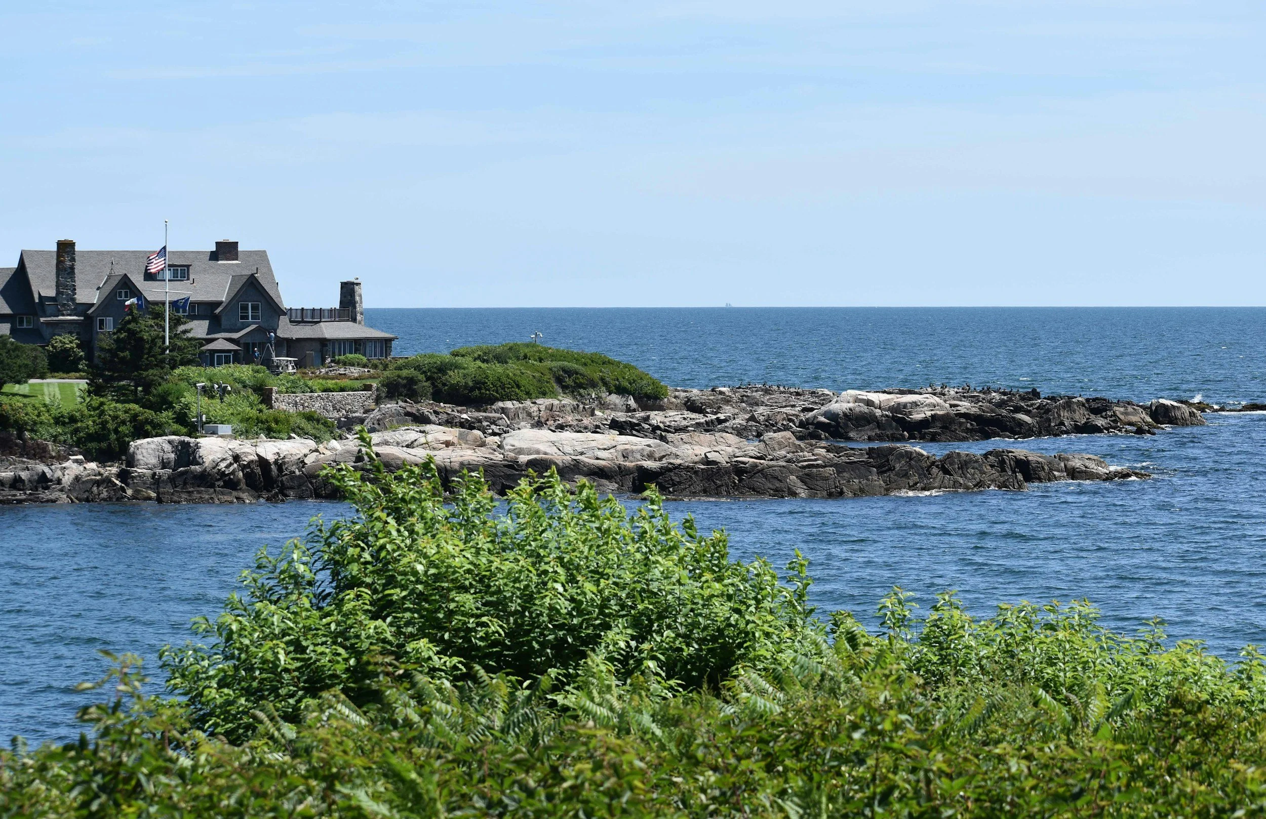A coastal house with gray walls and a gray roof situated on rocky terrain near the ocean, with lush green bushes and trees in the foreground and flags flying on a pole in front of the house.