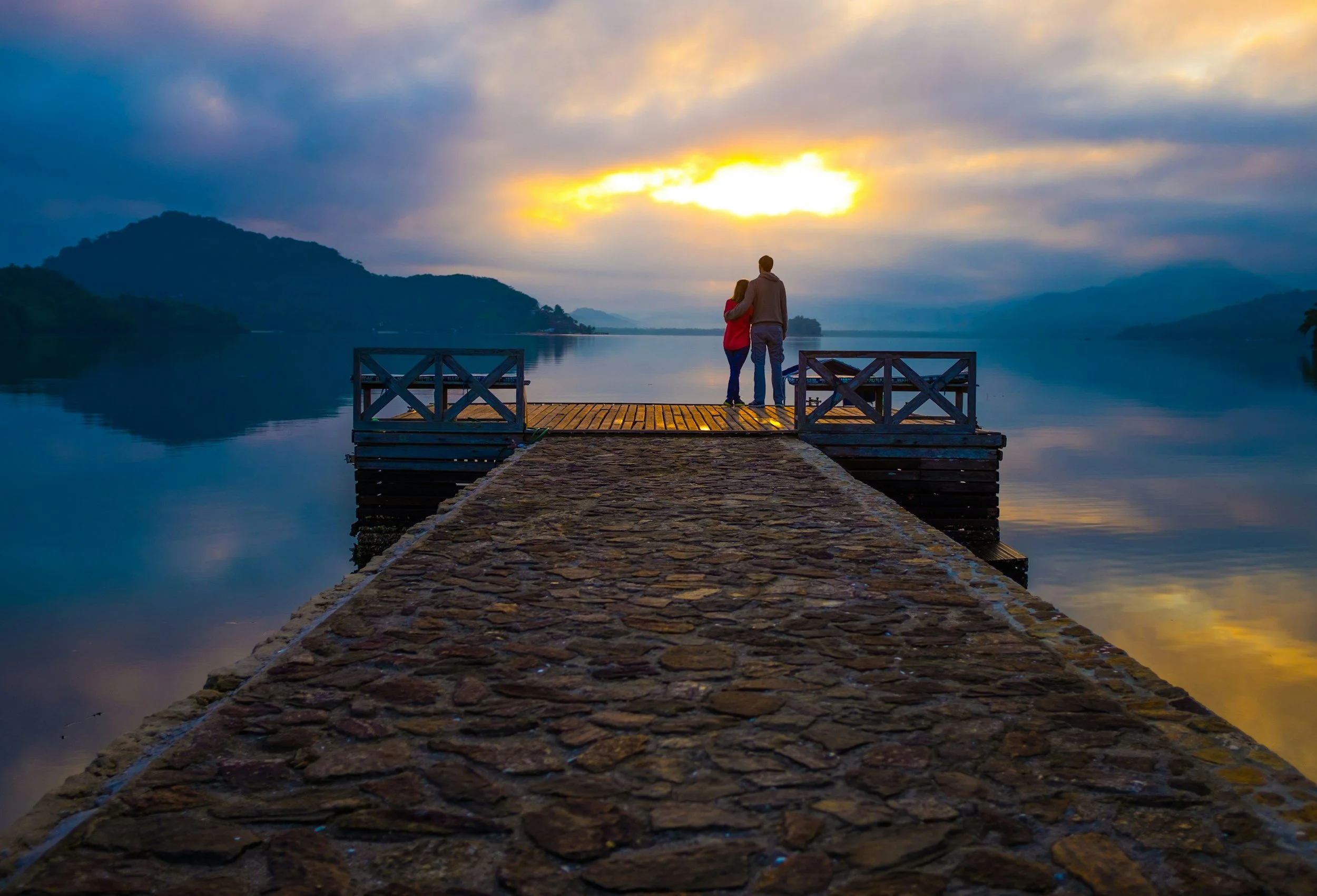 A couple stands on a wooden pier by a calm lake at sunset, with mountains in the background and clouds in the sky.