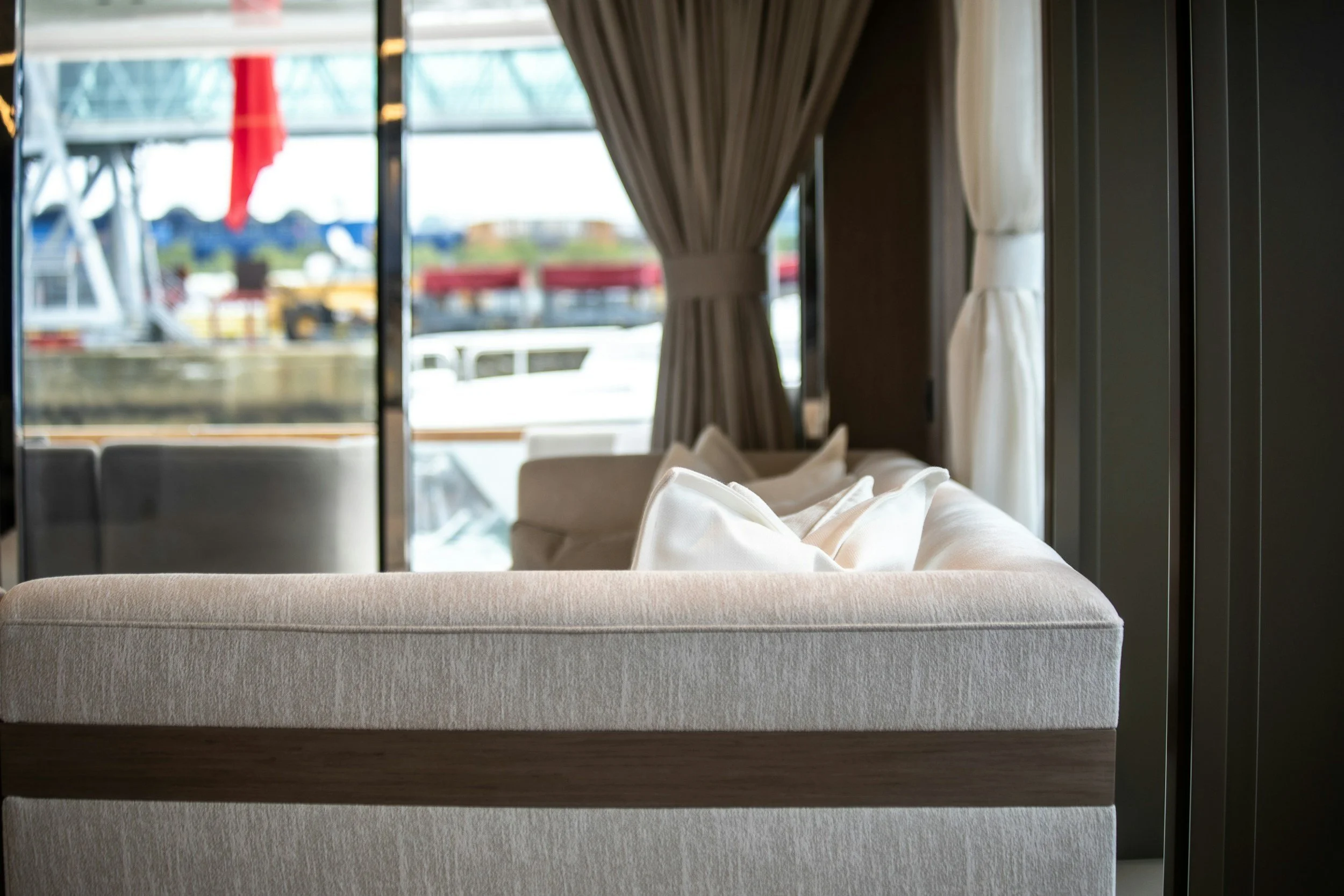 Interior of a modern living room with a beige sofa, decorative pillows, large windows with curtains, and a view of outdoor structures including a ferris wheel and red flags.