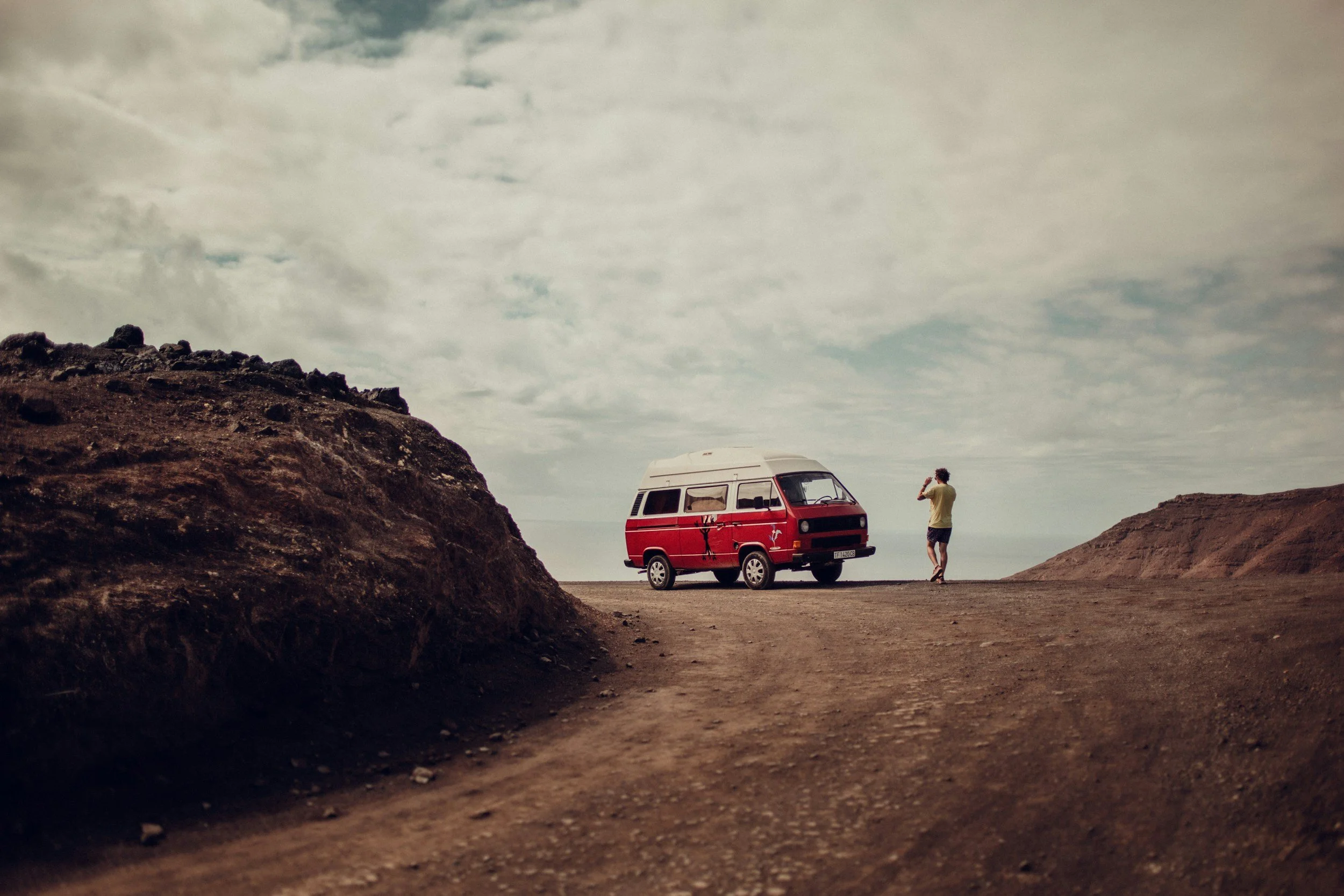 A person wearing shorts and a shirt standing next to a red van with a white top, on a flat open area with dirt ground. There are large rocks or mounds on both sides, and the sky is partly cloudy.