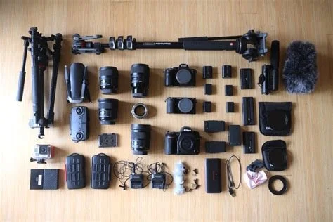 Various camera and audio equipment laid out on a wooden surface, including tripods, camera lenses, microphone, batteries, cables, and other accessories.