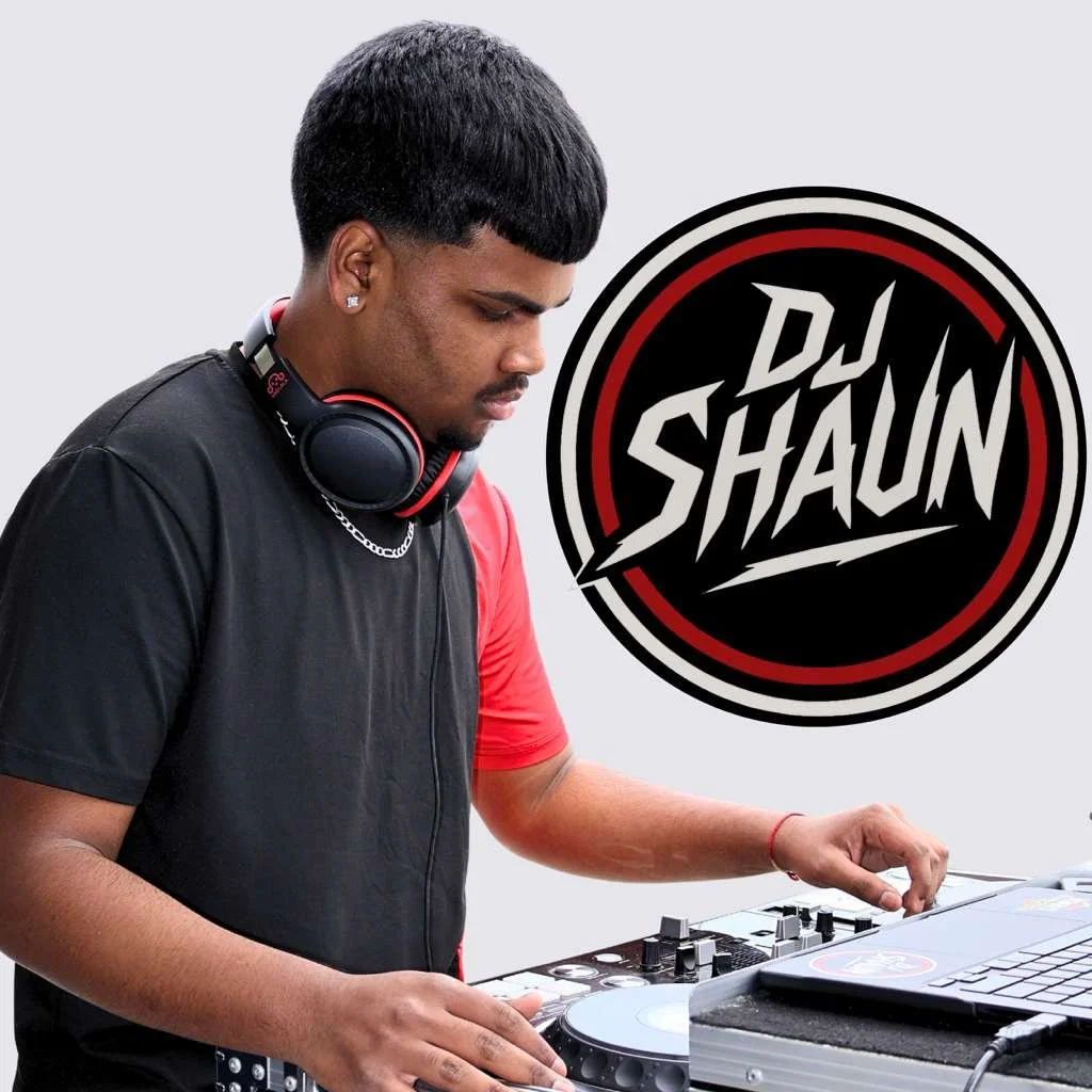 A male DJ with short dark hair, wearing a black shirt and headphones around his neck, standing at a DJ controller with a laptop nearby, with a DW Shaun logo in the background.