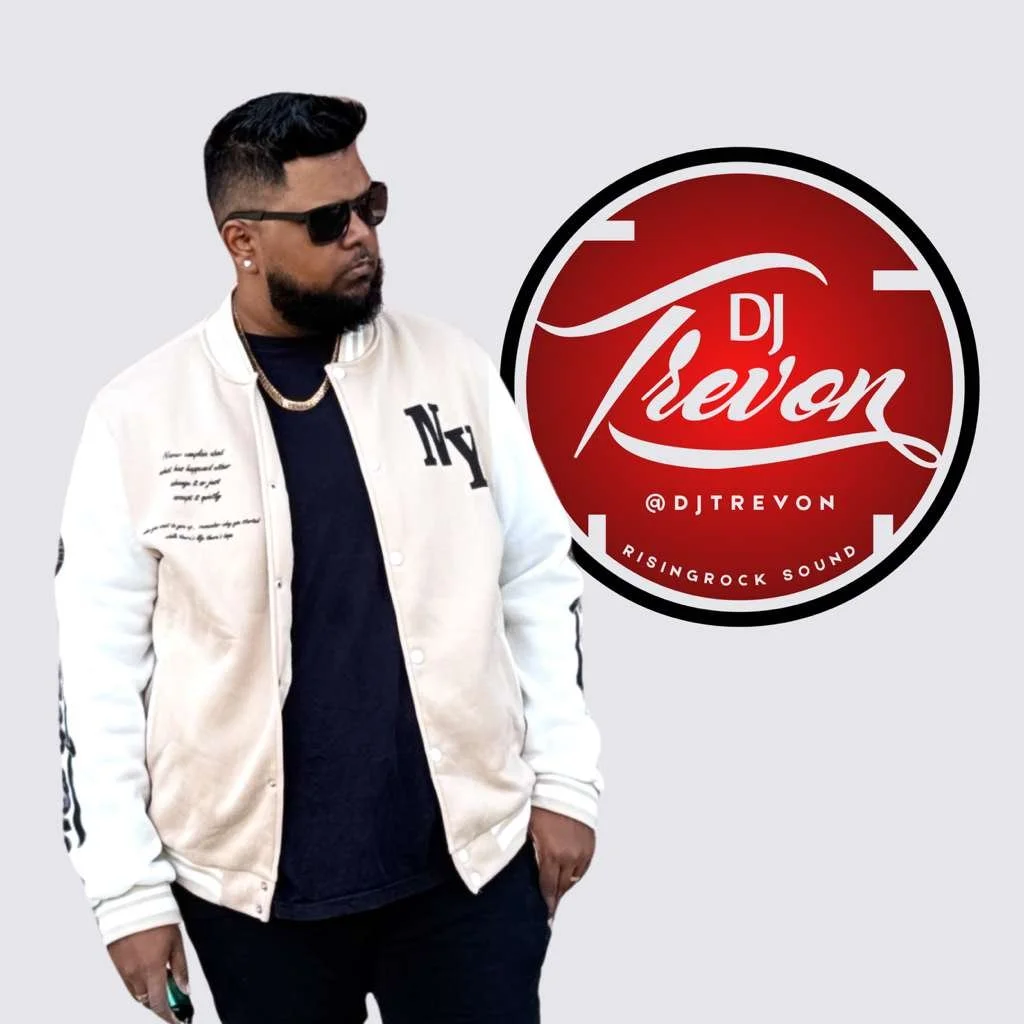 A man with dark hair and beard, wearing sunglasses, a white jacket with black lettering, and black clothing, standing next to a red circular logo that reads 'DJ Trevon @DJTrevon Rising Rock Sound.'