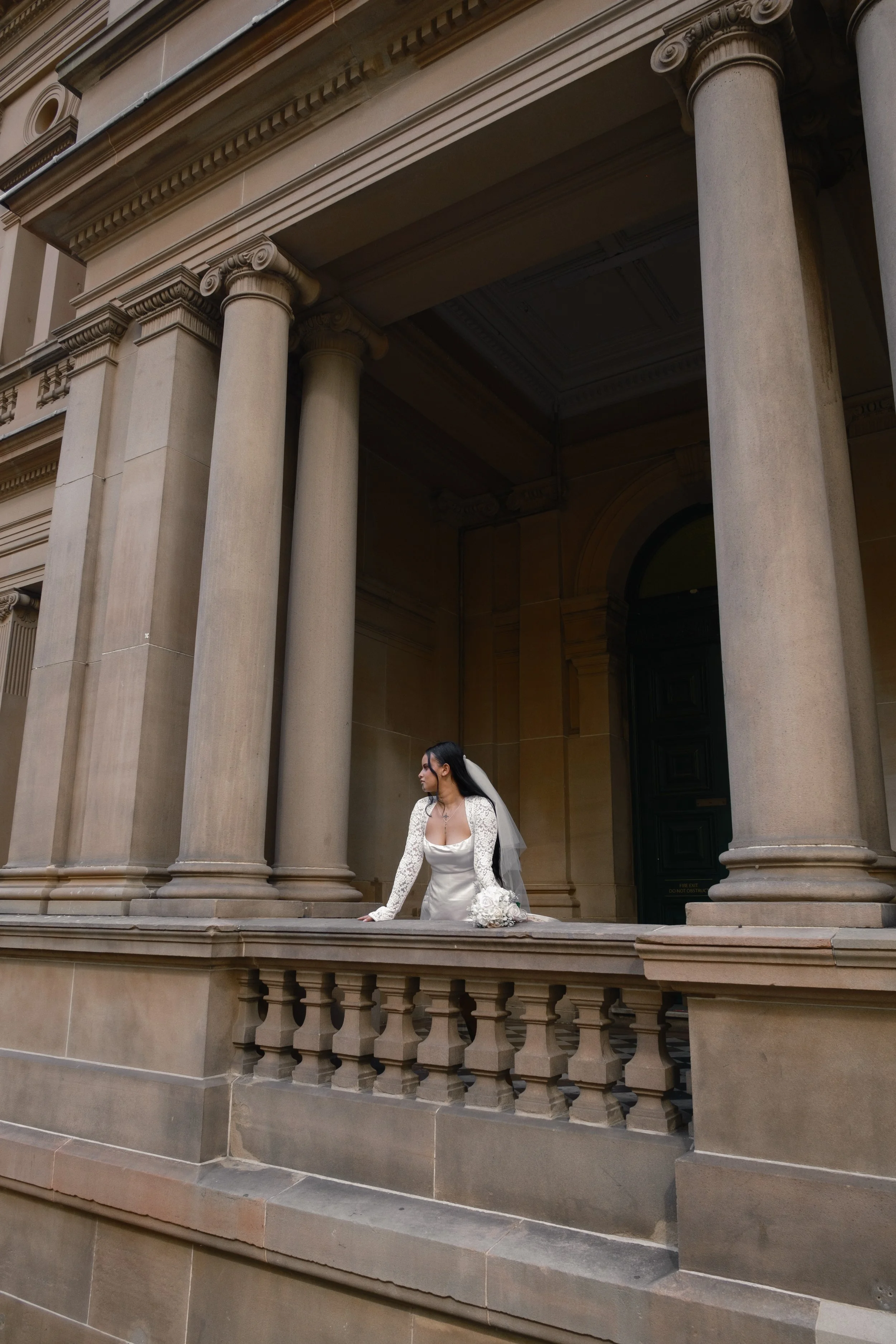A bride in a white wedding dress with long sleeves and lace details, holding a bouquet of white flowers, standing on the steps of a historic building with classical columns and ornate architectural details.