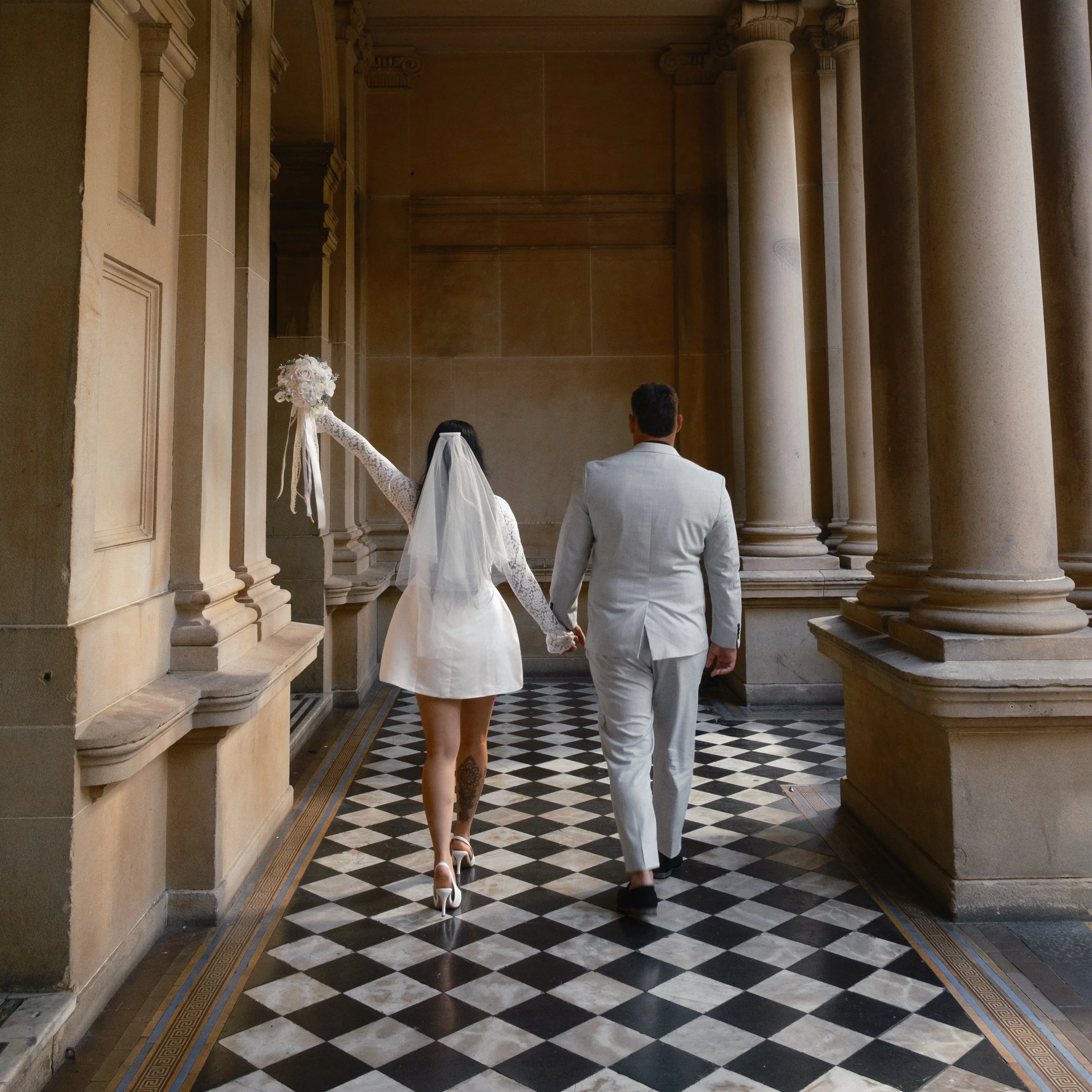 A bride and groom hold hands as they walk through an elegant, classical architectural corridor with tall columns and checkered black and white floor tiles.