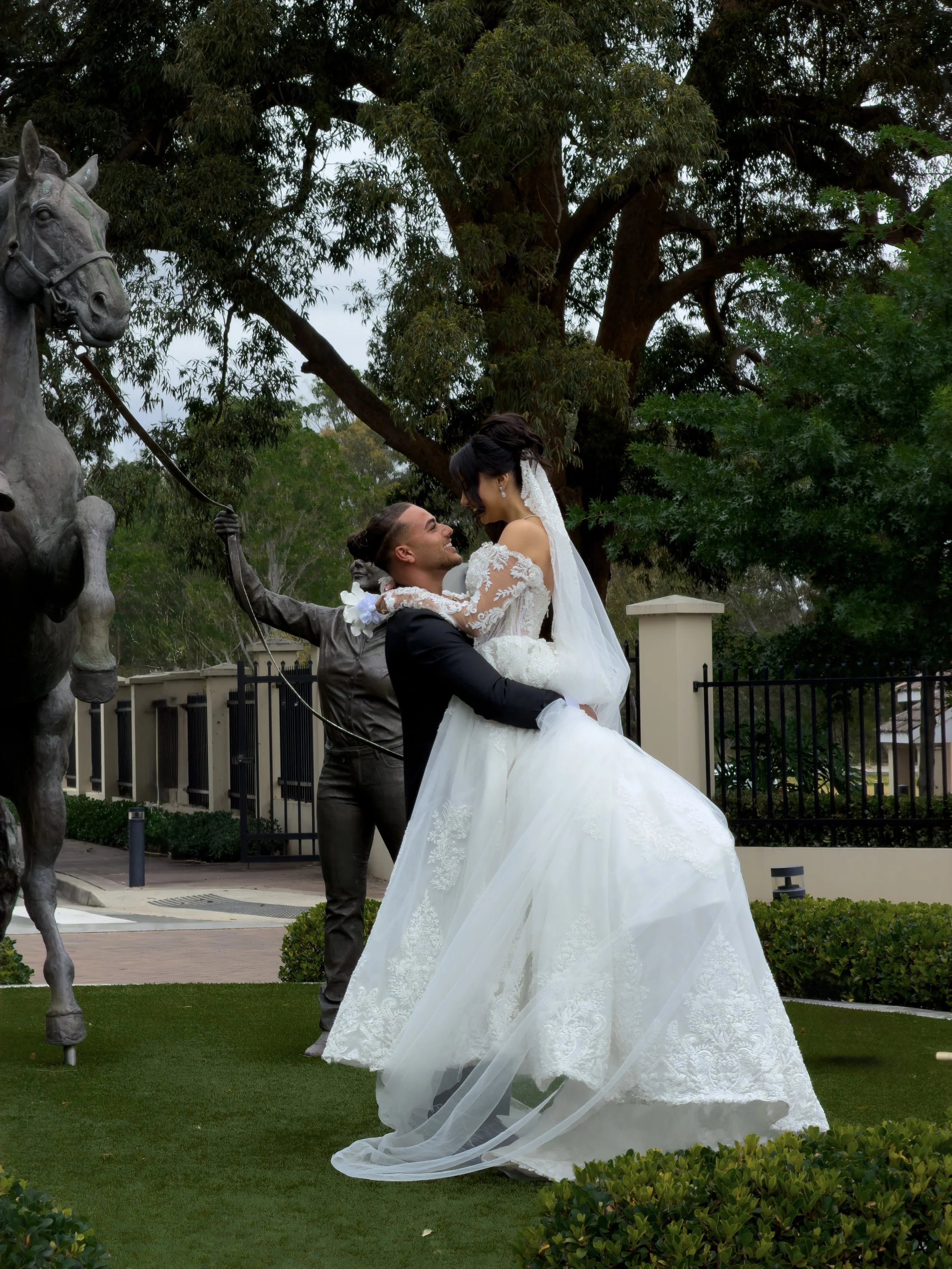A newlywed couple outdoors, with the groom lifting the bride, who is wearing a white wedding gown. They are smiling at each other under a large tree, with a statue of a horse on the left and greenery in the foreground.