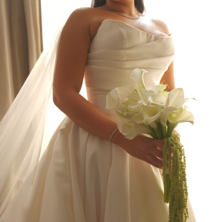 A bride in an ivory wedding dress holding a bouquet of white calla lilies and green hanging amaranthus, standing near a window with natural light.