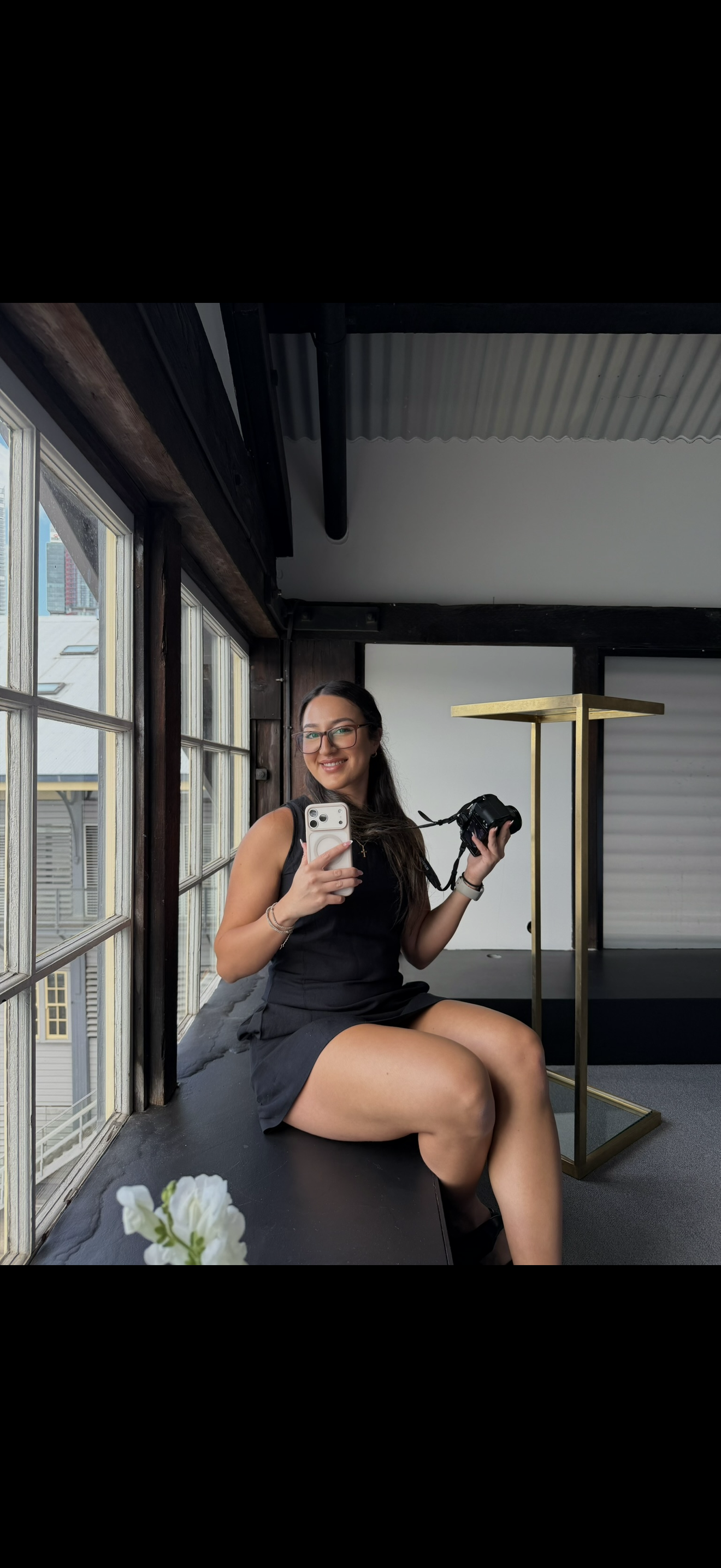 A woman with glasses and a black dress smiling while sitting on a windowsill, holding a smartphone in one hand and a camera in the other, with a cityscape visible through the window.