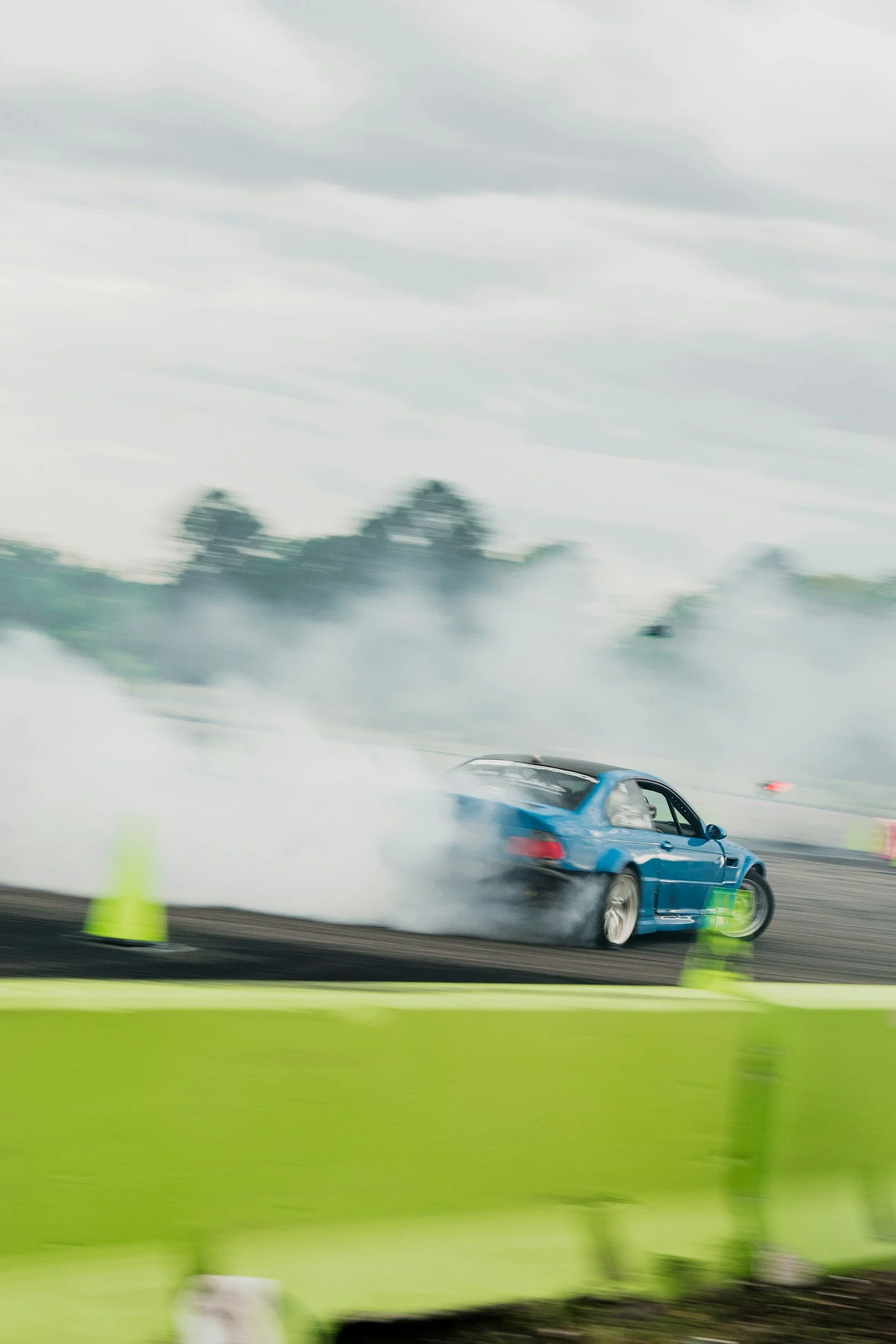 A Laguna Seca Blue E46 M3 car doing a burnout on a racetrack, producing a large cloud of smoke around the rear tires, with traffic cones nearby and a cloudy sky overhead.