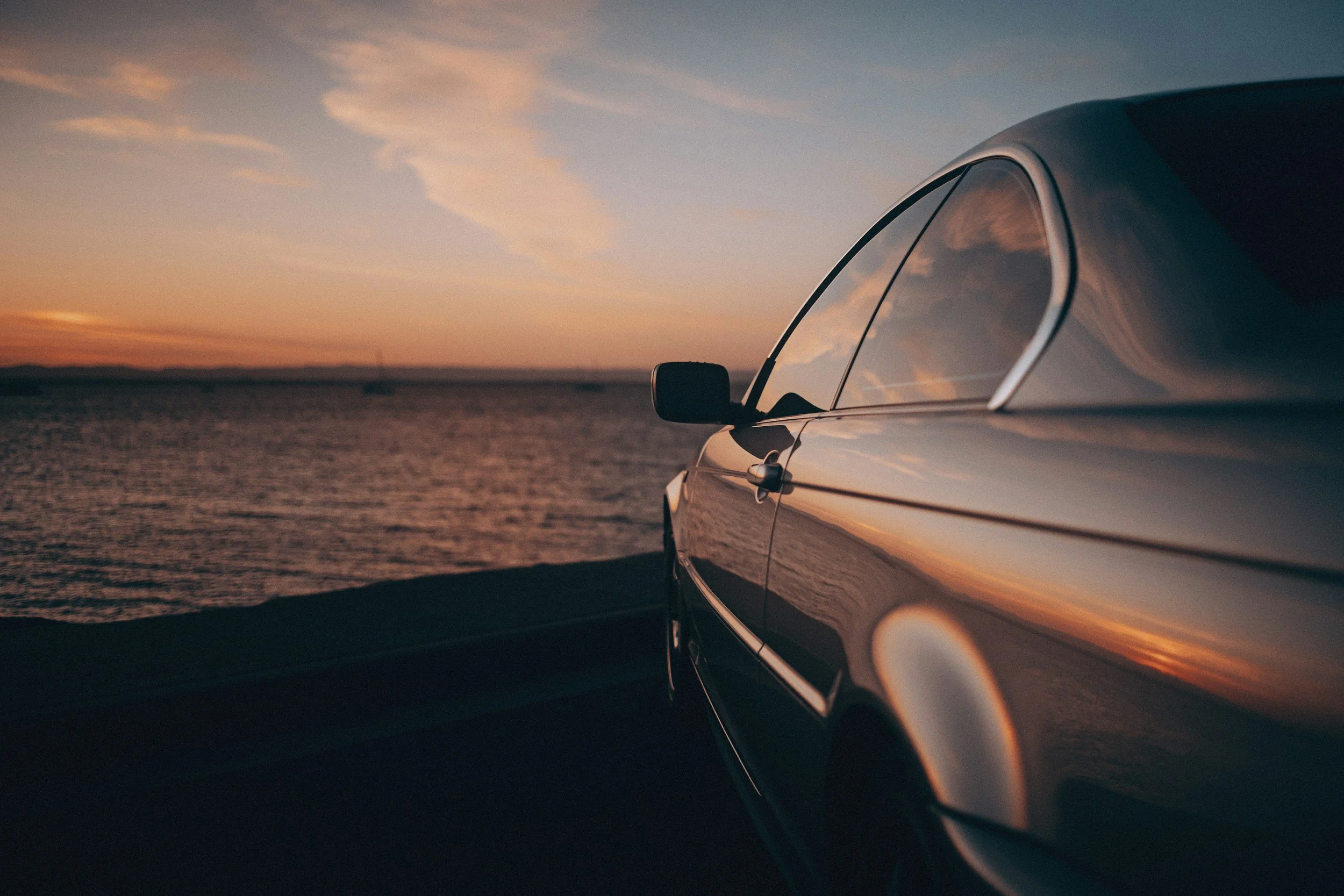 An E46 BMW parked near a body of water during sunset with the sky reflecting on the car's surface.