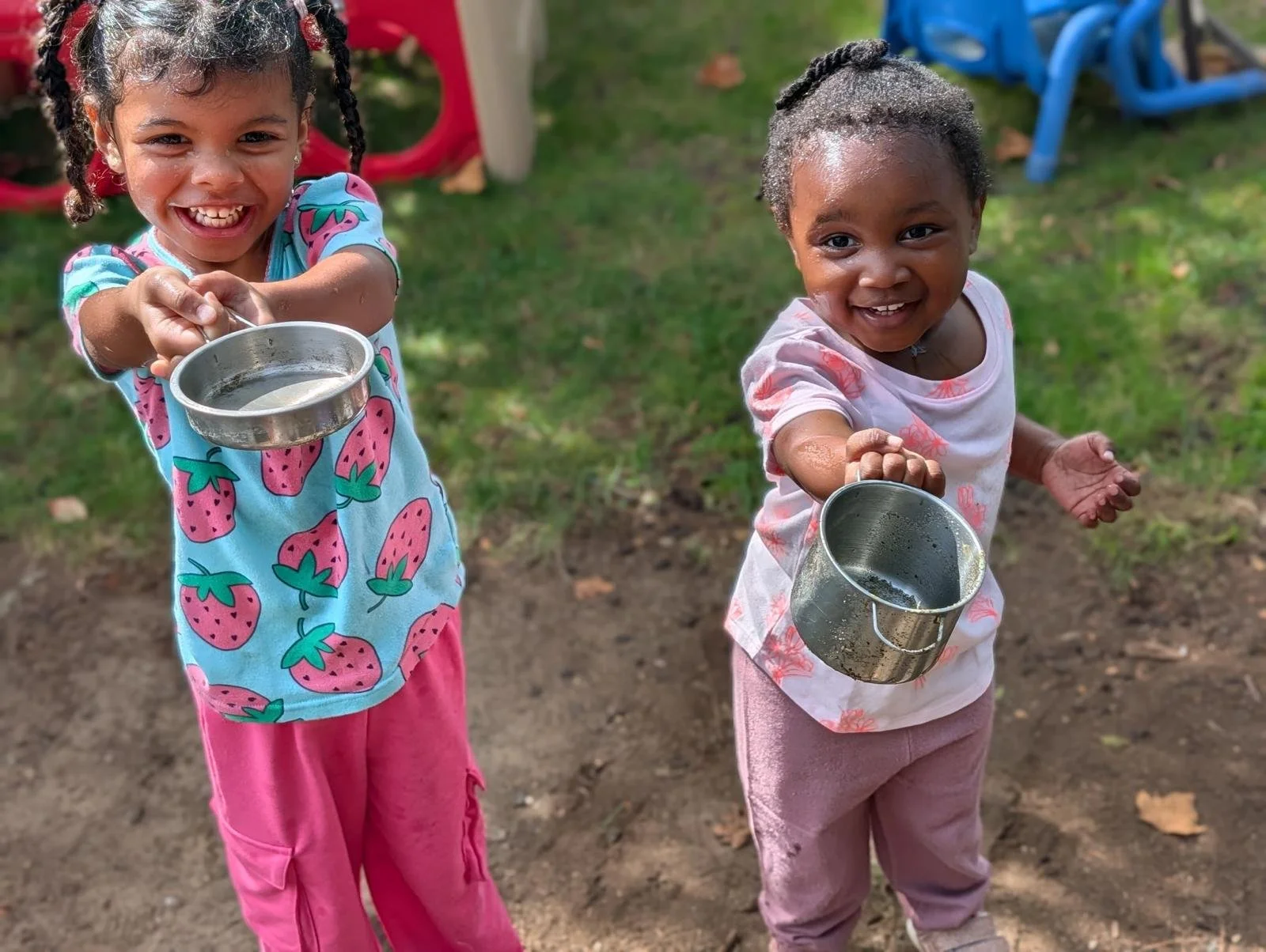 Two young girls smiling and holding metal cups outdoors on a grassy area with plastic chairs in the background.
