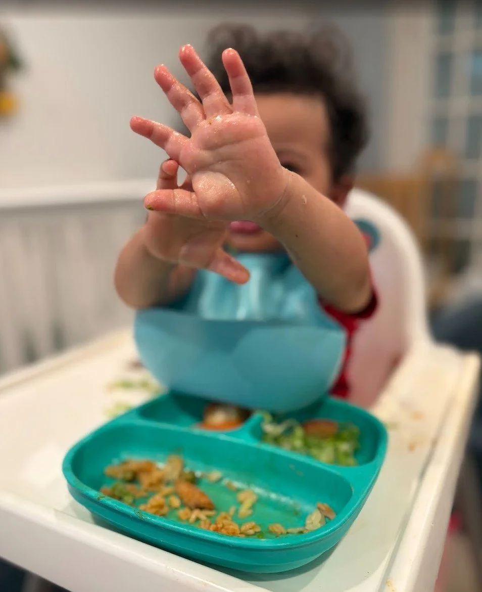 Child in a high chair reaching towards the camera, with food scattered in a divided turquoise plastic plate and a blue bowl, set on a white tray in a kitchen.