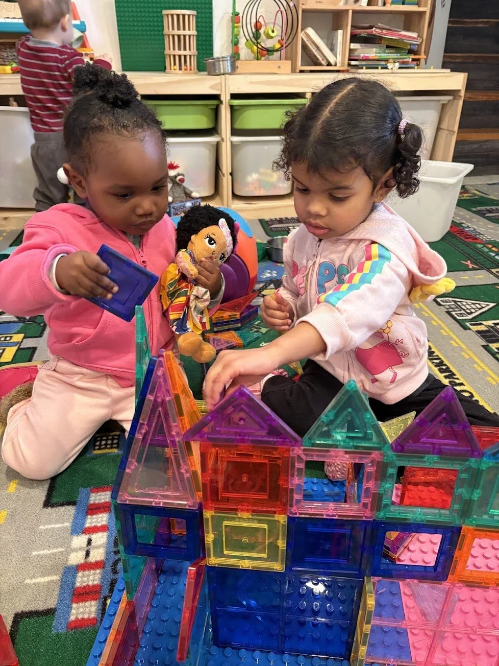 Two young girls playing with colorful magnetic building blocks in a classroom, with a third child in the background and shelves filled with toys and books.