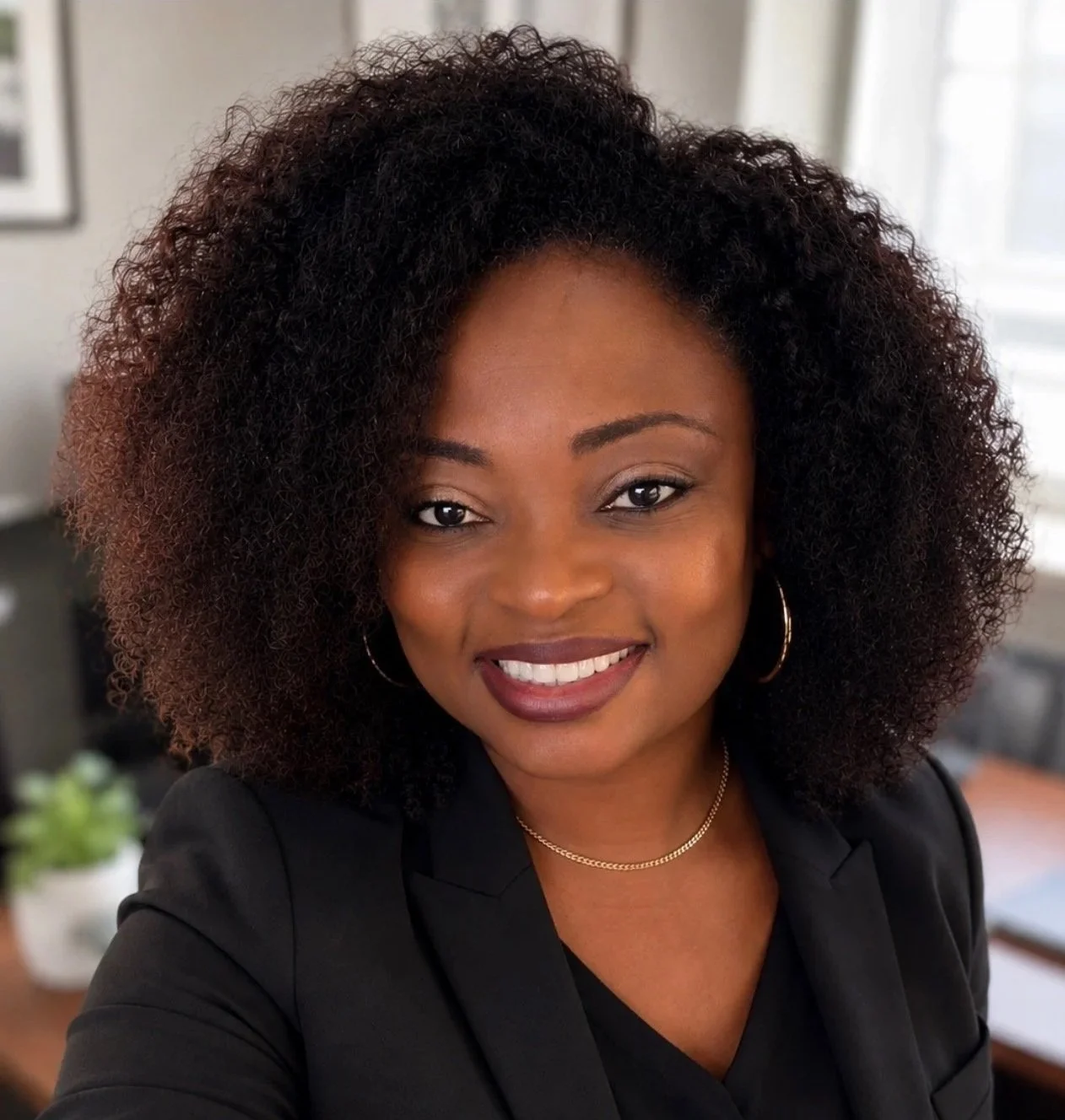Close-up of a smiling African American woman with curly hair, wearing a black blazer, hoop earrings, and a gold necklace, indoors with a blurred background.