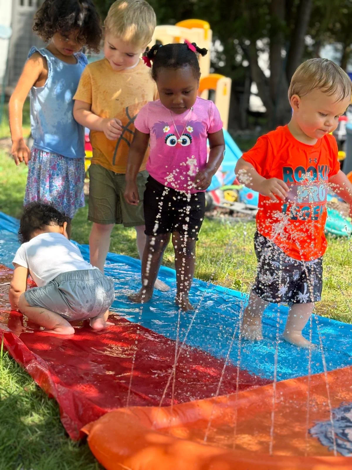 Children playing in a water game outdoors on a sunny day, with some standing and splashing water on a slip and slide float.