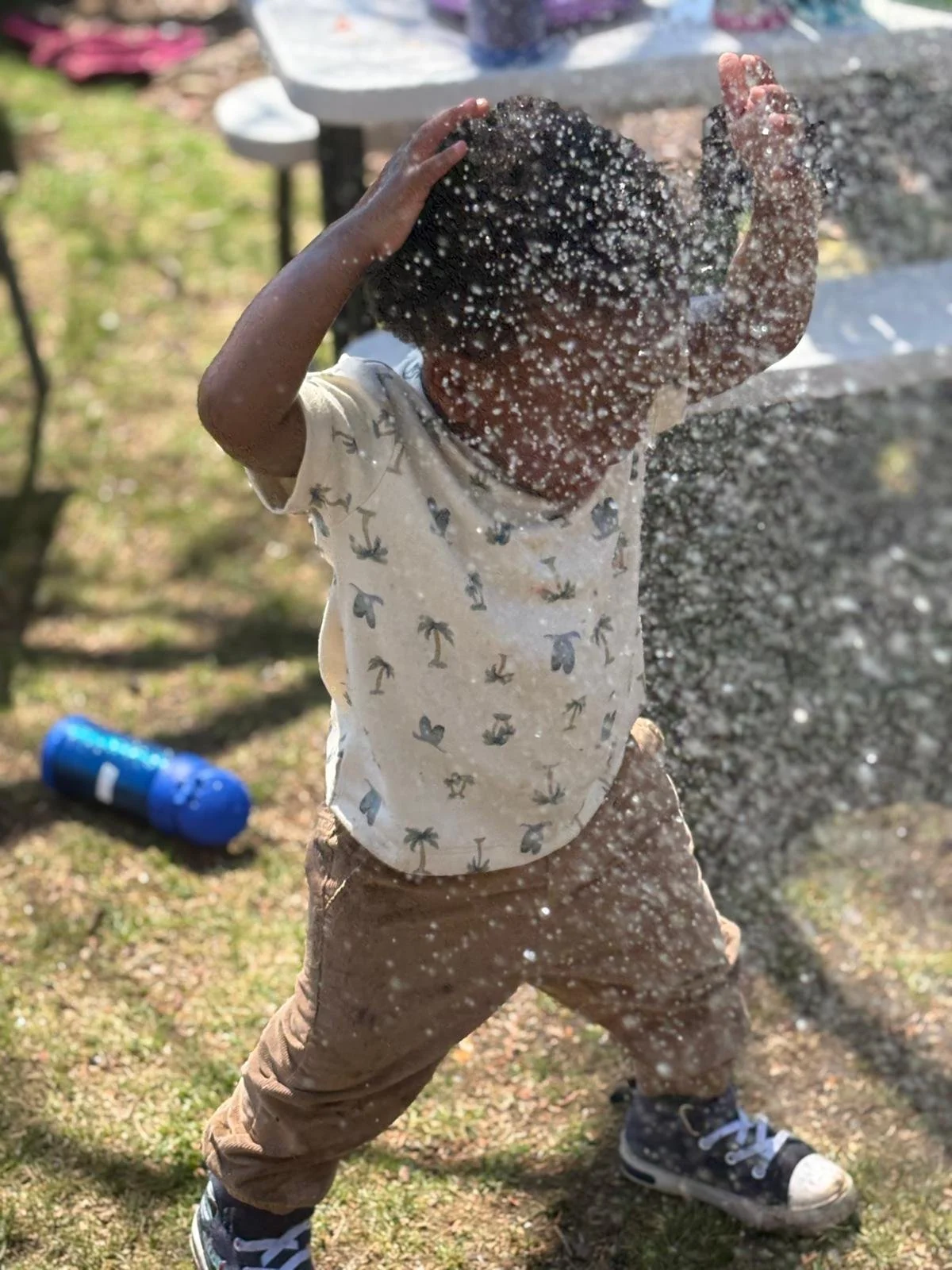 A young boy outside is playing with water, splashing his face with his hands. The water droplets are visible in mid-air around his head. He is wearing a beige t-shirt with palm tree prints, tan pants, and black sneakers with white toes. There is a blue water bottle on the grass behind him, and a picnic table with a white object on it in the background.