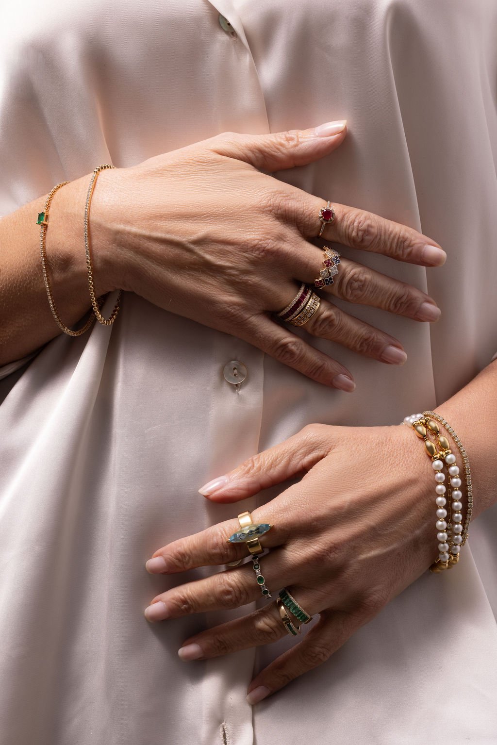 Manos con varios anillos y pulseras, descansando sobre una blusa de seda beige.
