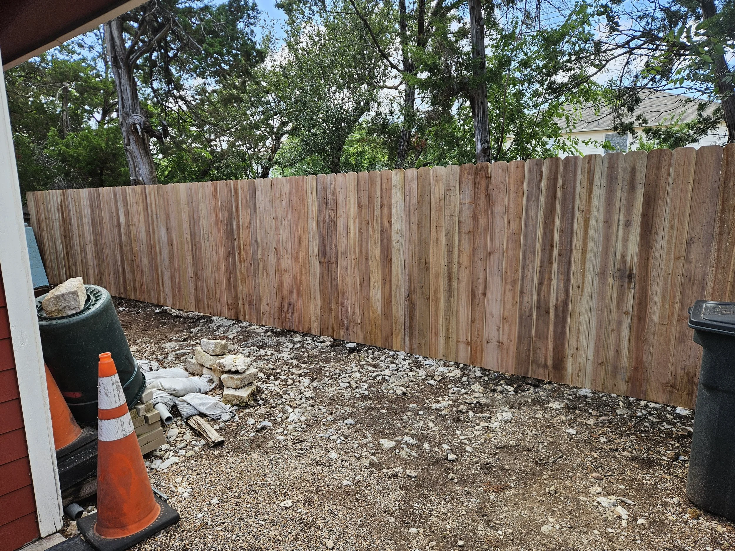 A backyard with a wooden privacy fence and trees in the background. Construction materials and orange cones are in the foreground, indicating ongoing outdoor work.