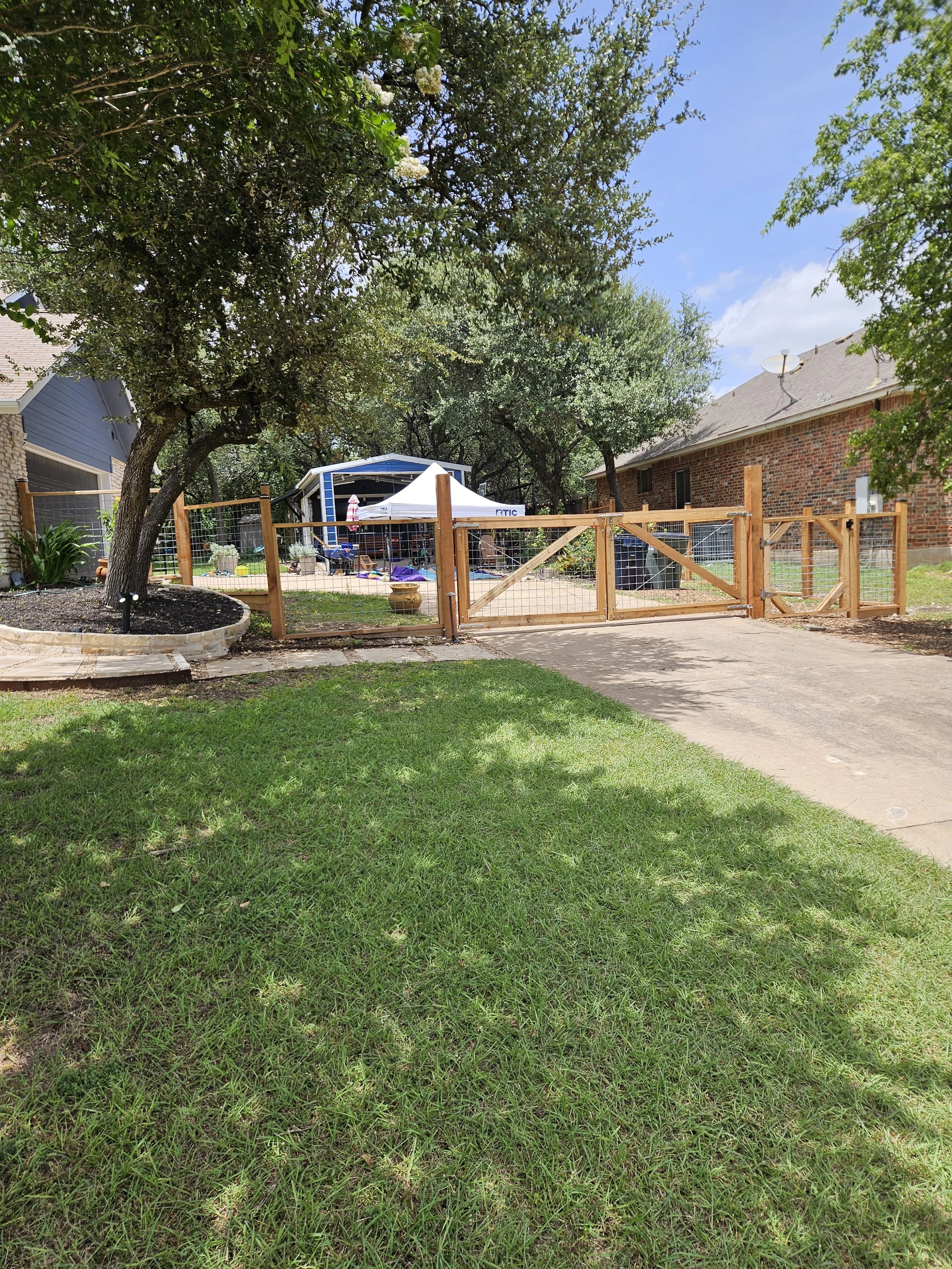 A backyard scene with trees, a brown wooden fence, a concrete walkway, and a small shed with a white canopy and gardening tools underneath. The sky is partly cloudy with some blue visible.