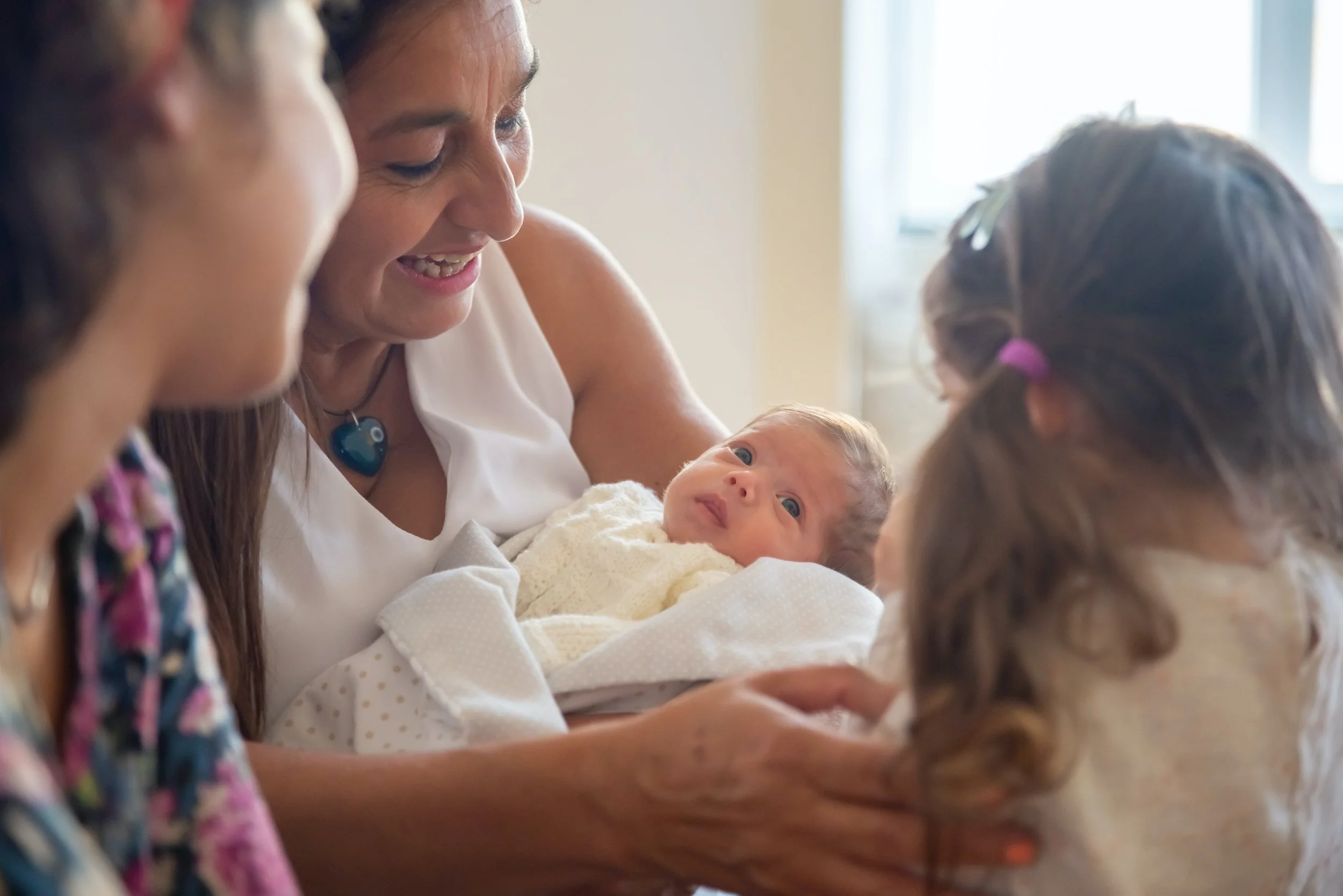 A woman holding a newborn baby in a white outfit, smiling and interacting with two young girls around her.