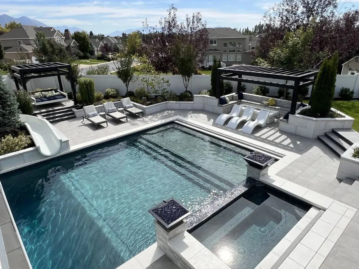 A backyard with a swimming pool, poolside lounge chairs, and two shaded seating areas with beds and chairs, surrounded by trees and a white fence.