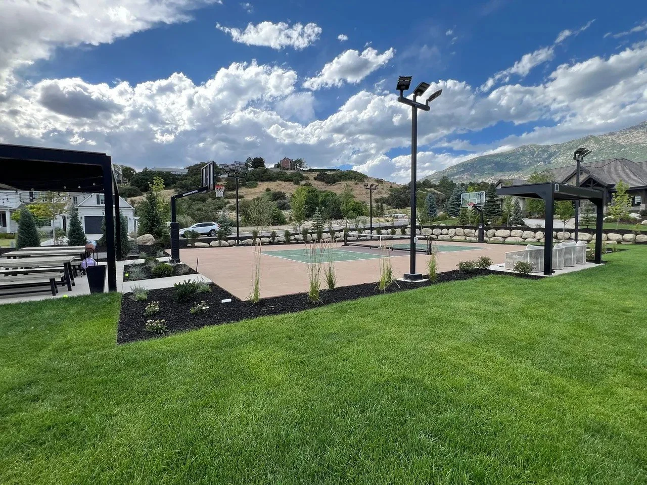 A recreational outdoor sports court with basketball hoops, surrounded by green grass, with some outdoor seating and a landscaped garden area, set against a backdrop of hills and a partly cloudy sky.