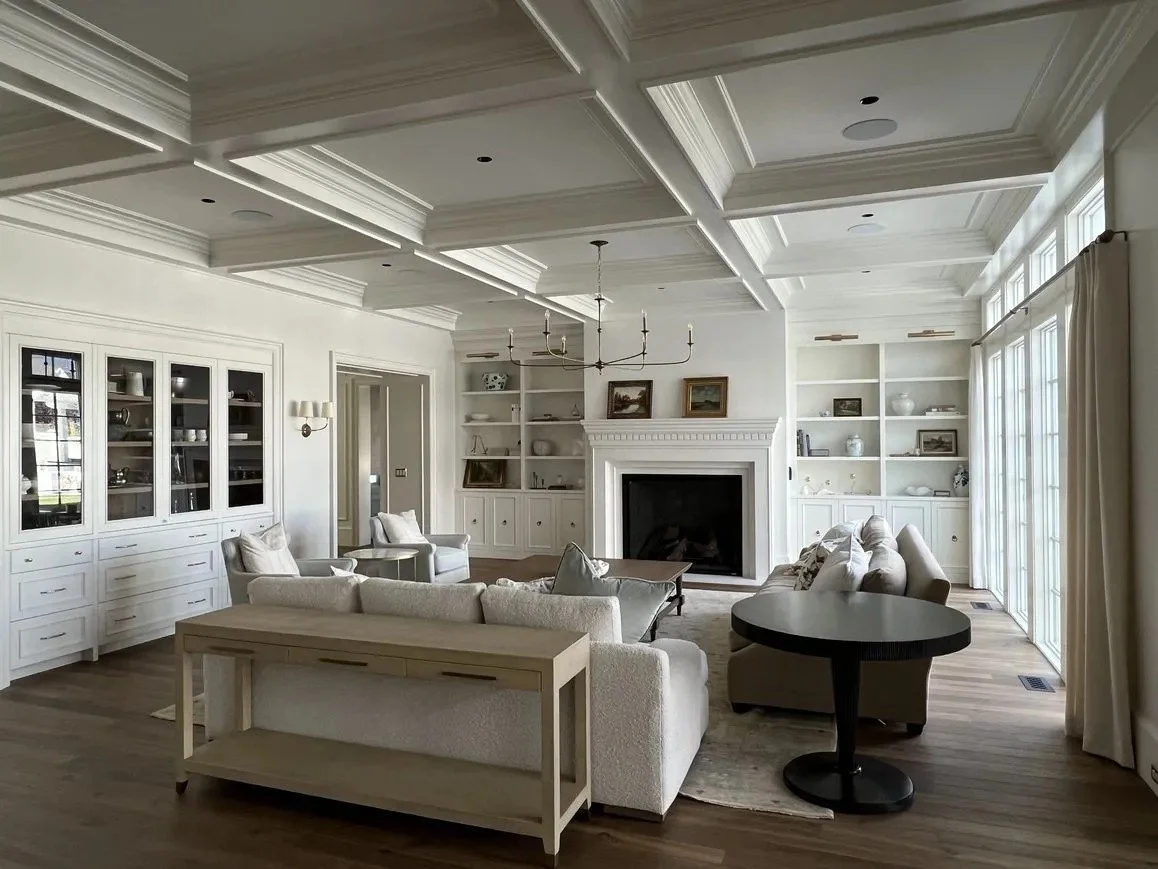 Bright living room with white walls, built-in shelves, a fireplace, a large window with curtains, and white and beige furniture.