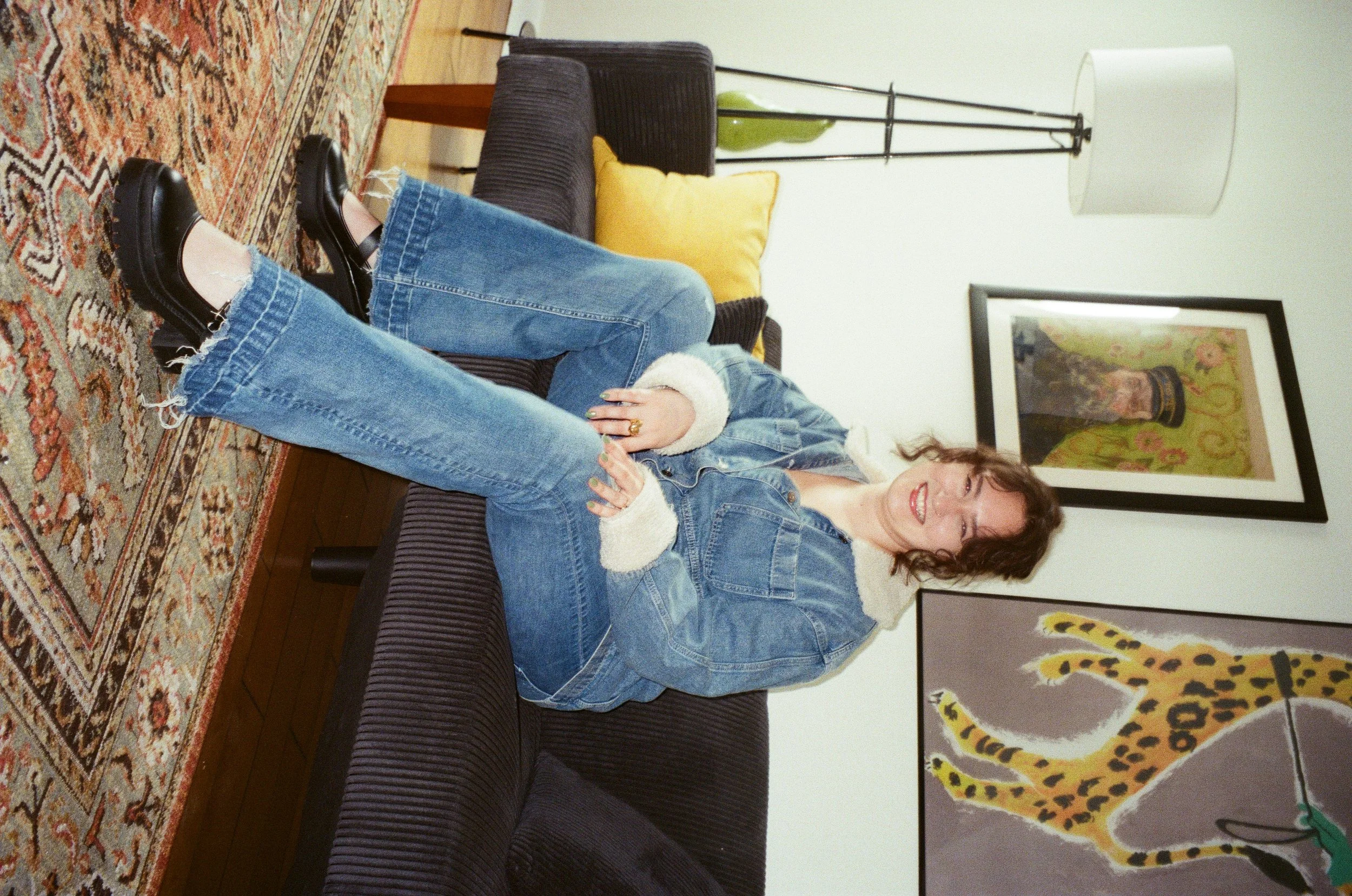 Brunette woman wearing a jean jacket and jean pants and black shoes sits on a black couch smiling.