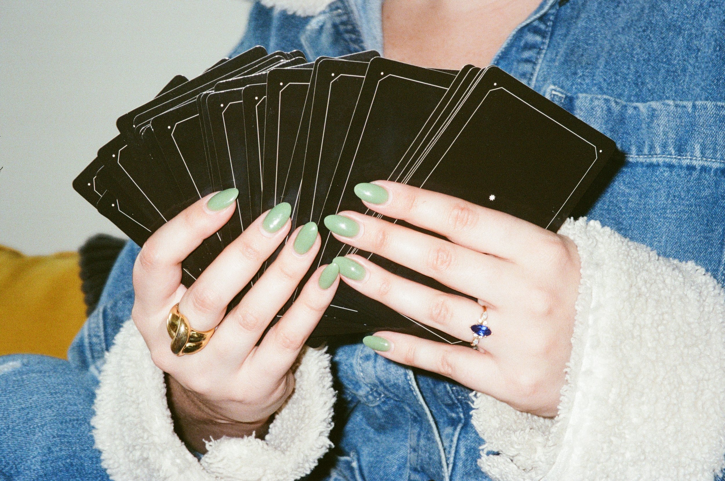 A person holding a fan of black cards with white outlines on the back. The person has green nail polish and gold and silver rings, and is wearing a denim jacket and white sweater.