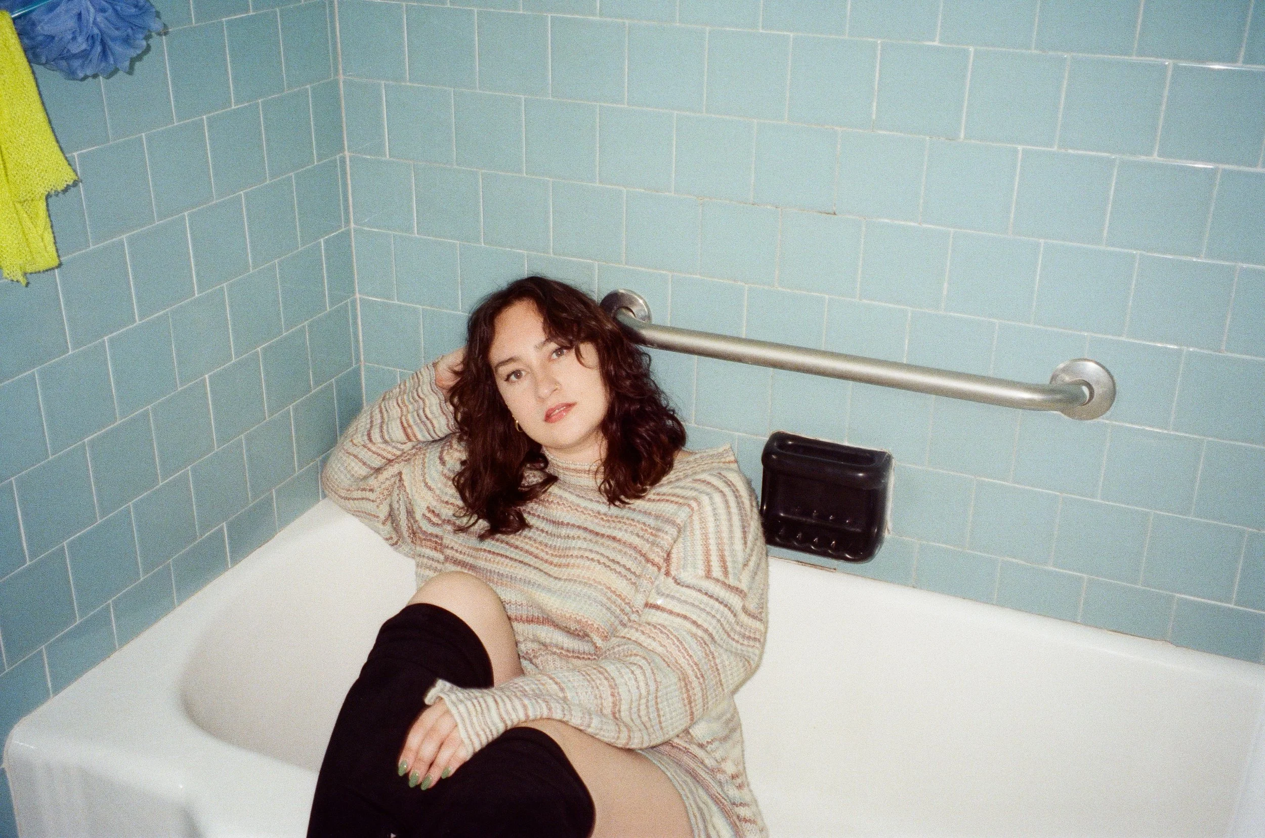 Brunette woman wearing a sweater and black boots sits in a bathtub with blue tile
